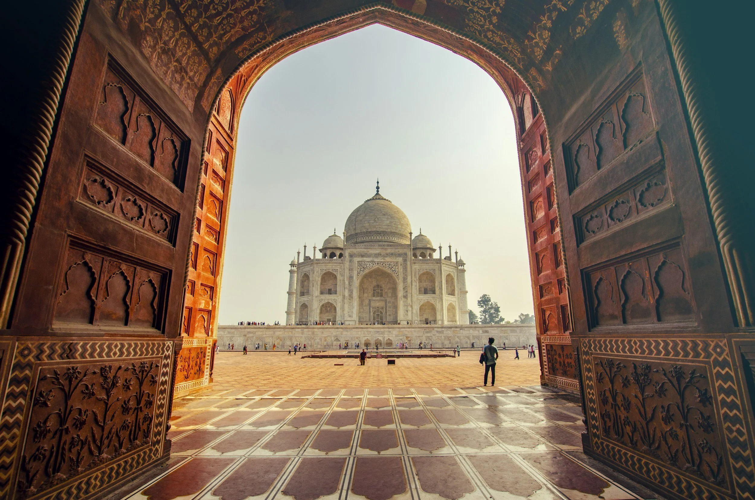 View of the Taj Mahal through a large, ornately decorated entrance door with intricate woodwork and carvings, showing the white marble monument in the distance against a clear sky.