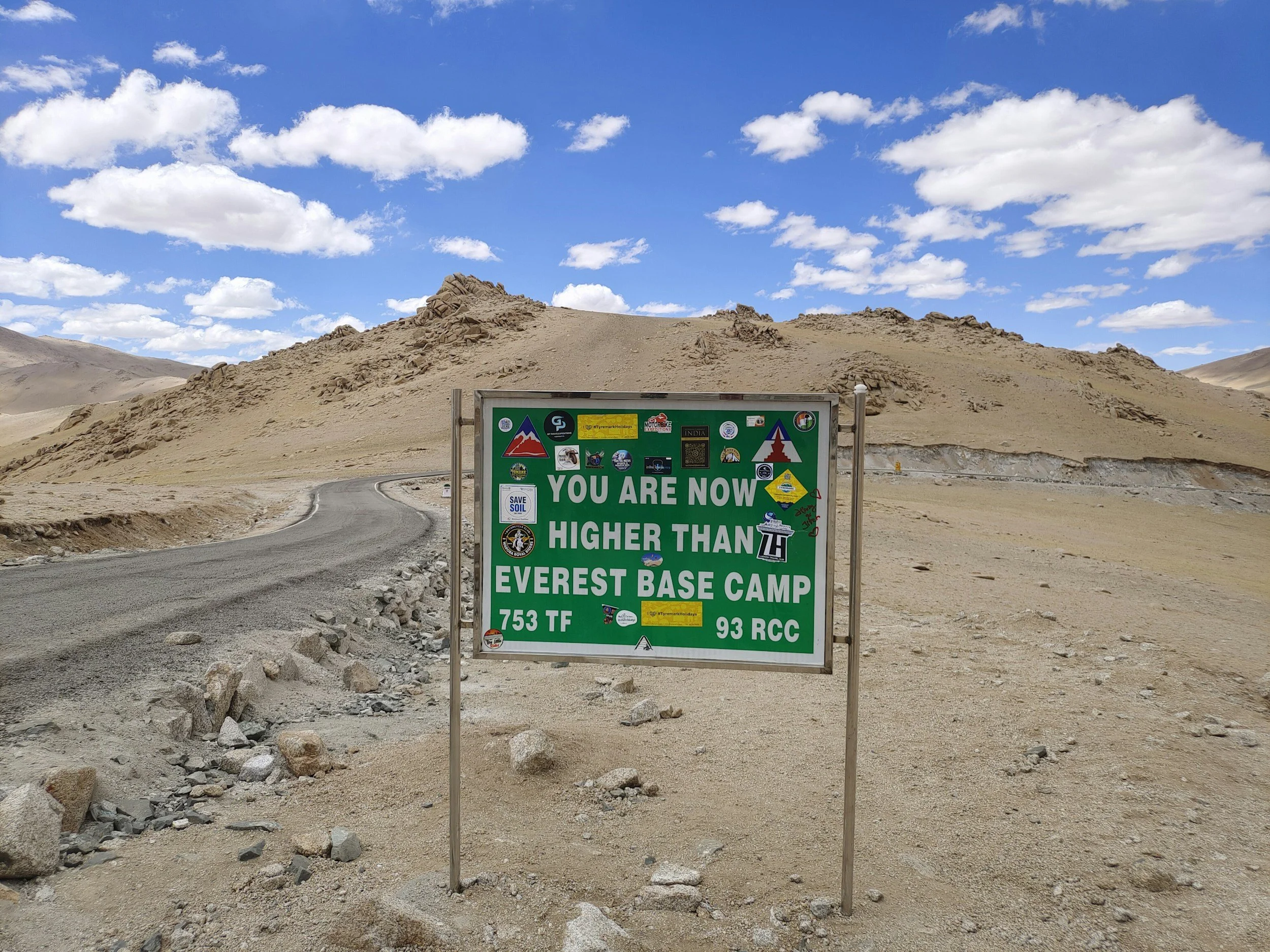 A sign in a barren desert landscape at Everest Base Camp indicating the elevation of 7535 meters, with a winding dirt road and rocky hills in the background under a partly cloudy blue sky.