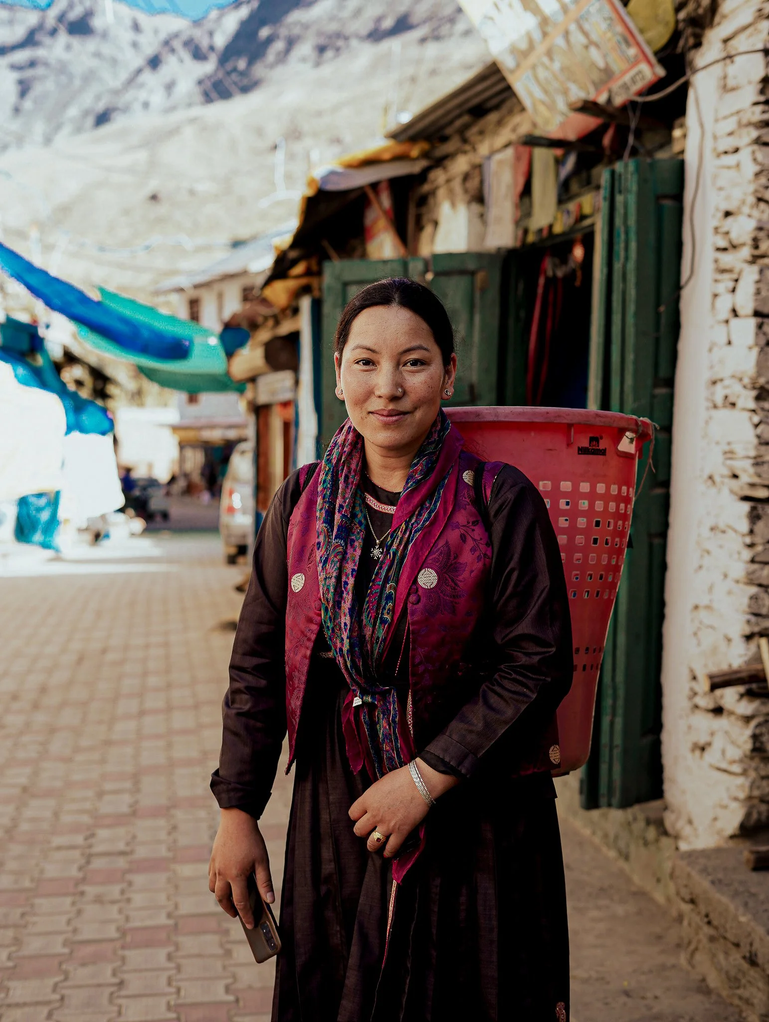 A woman standing on a paved street in a mountainous area, wearing a dark dress and colorful scarf, with a large red basket on her back, holding a phone in her left hand.