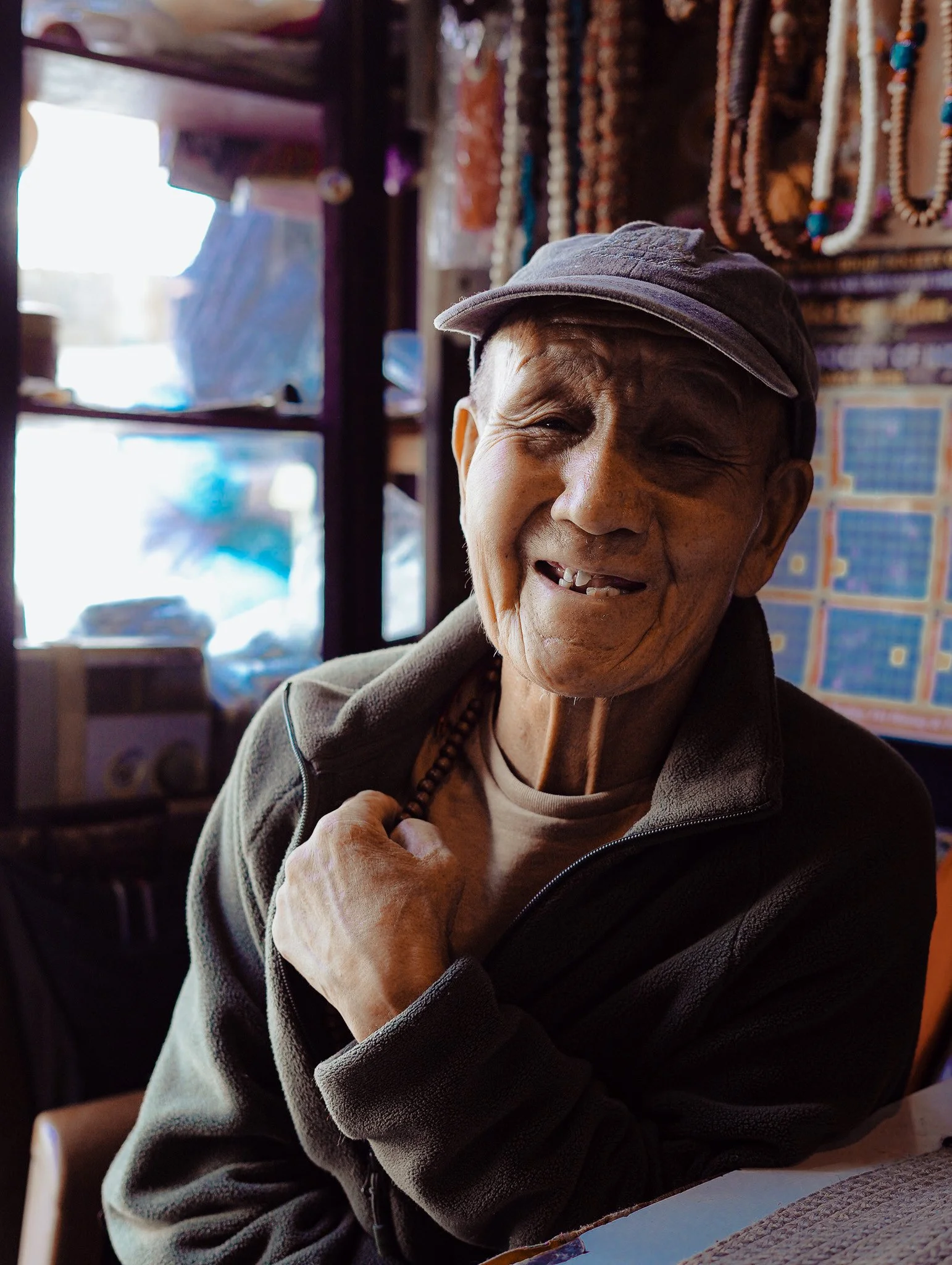 An elderly Indian man smiling and wearing a cap, inside a shop with colourful necklaces and posters in the background in Leh during our Himalayan Motorcycle adventure