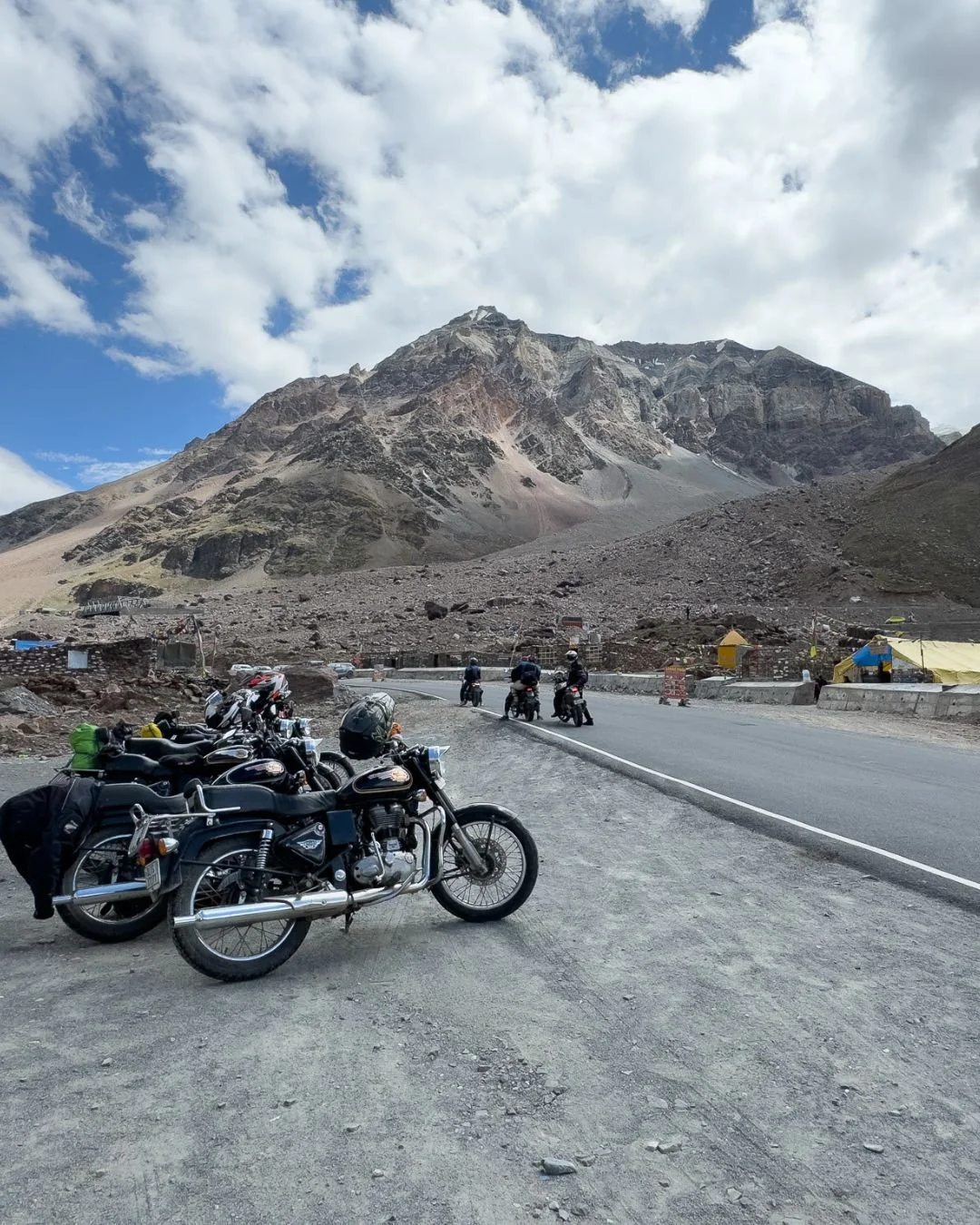 Motorcycles parked by a mountain road in the Himalaya in India with bikers riding motorcycles in the background, mountain peak with rocky terrain and partly cloudy sky.