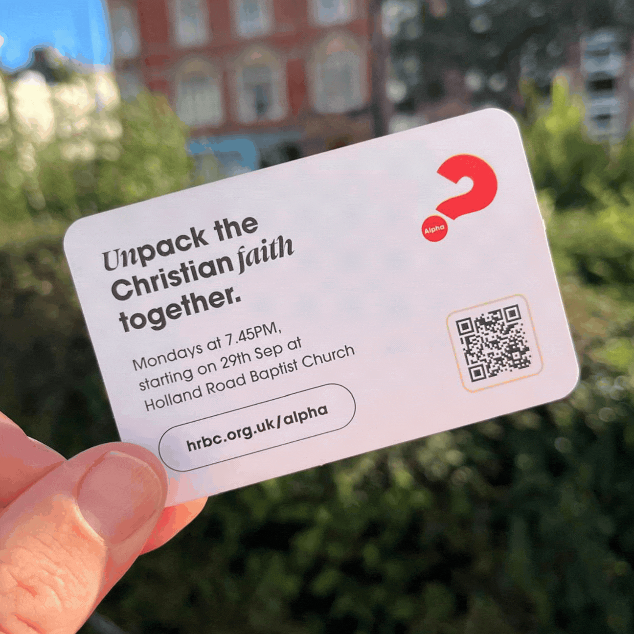 A person holding a card outdoors against a background of trees and buildings. 