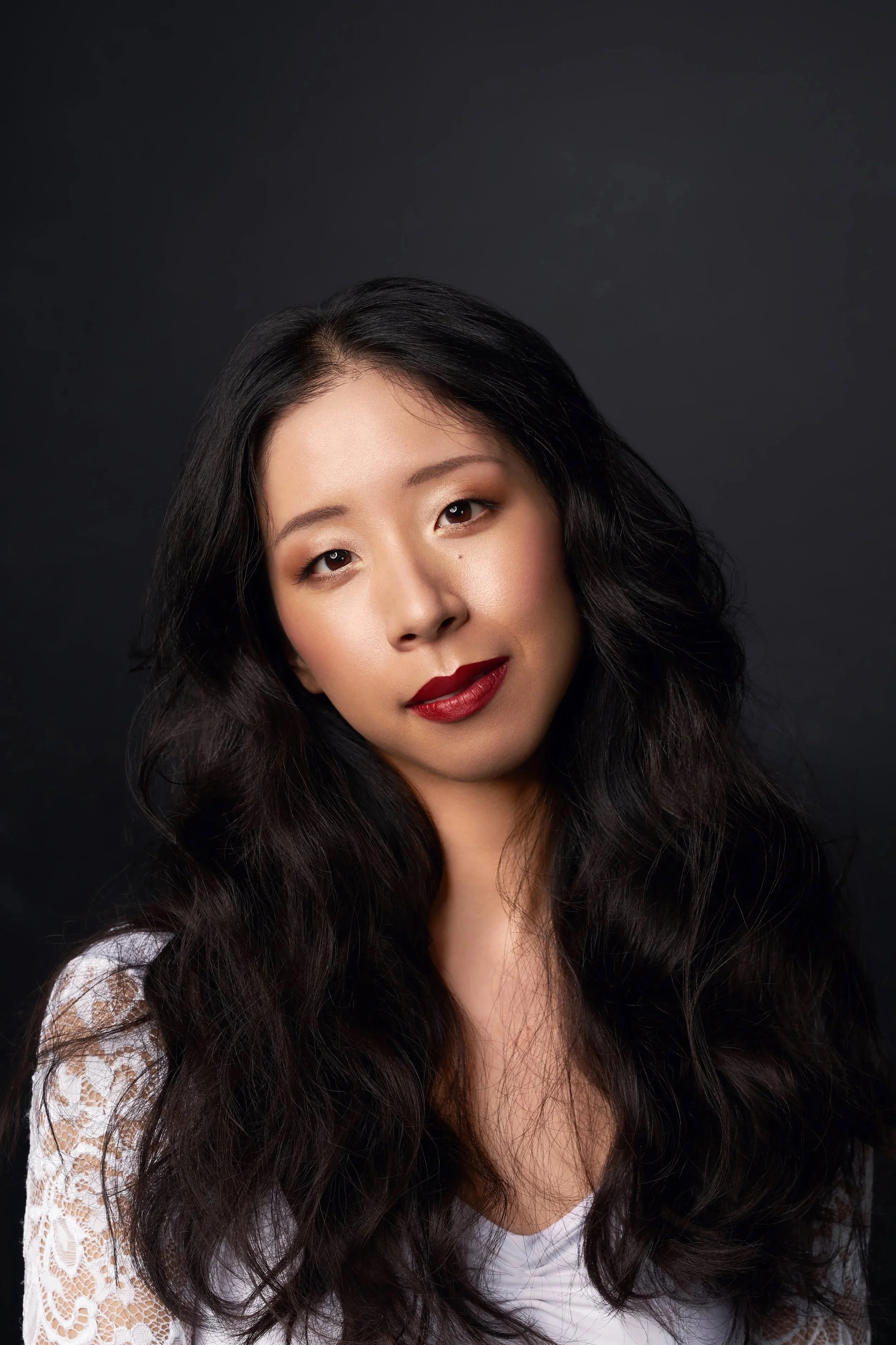 A portrait of an Asian woman with long, wavy black hair, wearing makeup with dark lipstick, and a white lace top, against a dark background.
