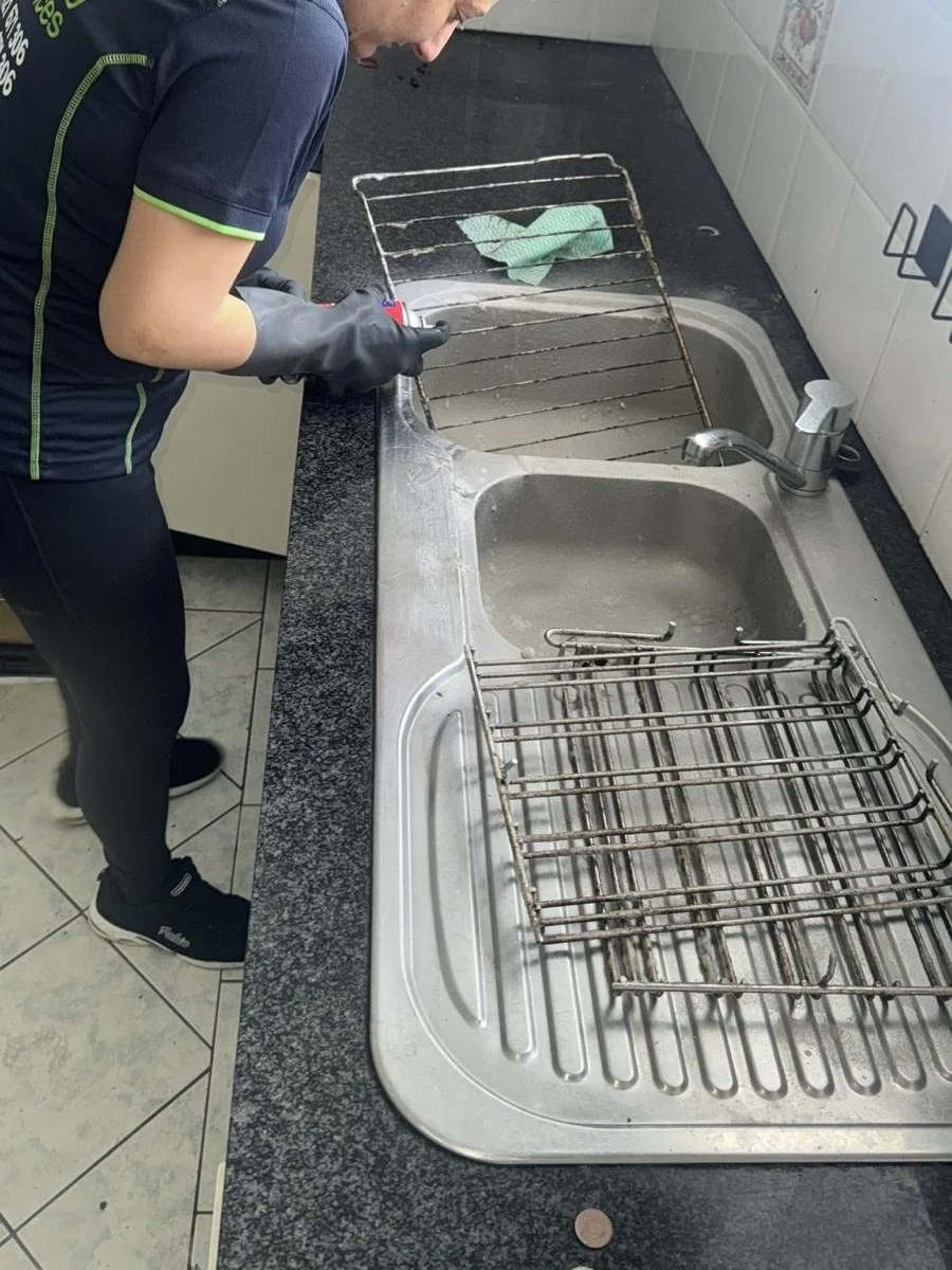 Person wearing black clothes and gloves standing in a kitchen, holding a wrench near a rusty metal grill rack over a large kitchen sink with a stainless steel dish rack on one side. A green cloth is on the metal grill rack.