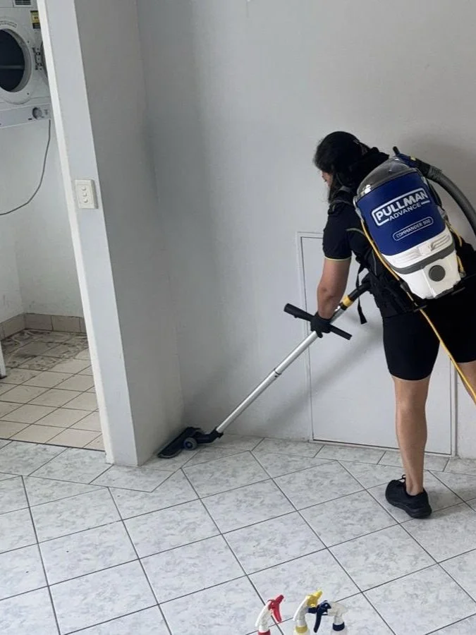 A person wearing black clothing and a backpack vacuum cleaner is cleaning the tiled floor near a wall, in a room that appears to be a laundry area with a washing machine in the background.