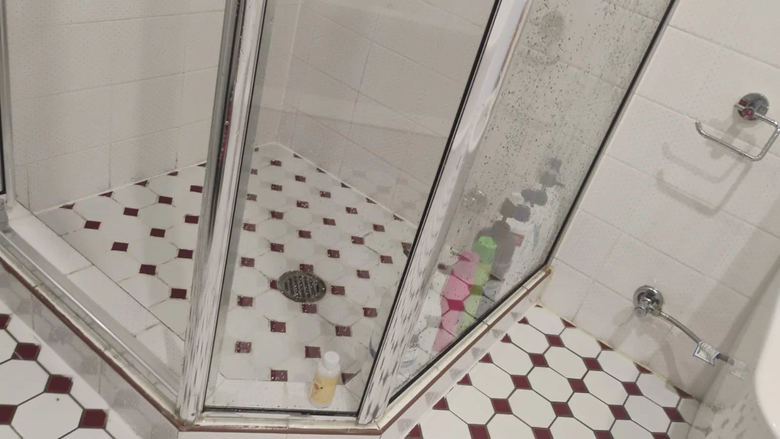 A bathroom shower area with a glass enclosure, white tiles with red diamond accents on the floor, a drain in the center, and various bottles inside the shower. The wall tiles are white with a subtle pattern.