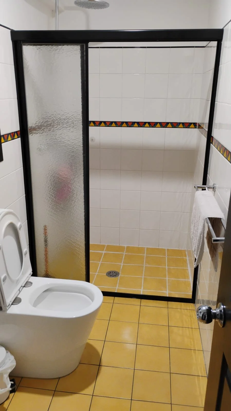 Bathroom with a toilet in the foreground, a glass shower enclosure with a black frame, white tiled walls with a colorful decorative border, and yellow tile flooring.