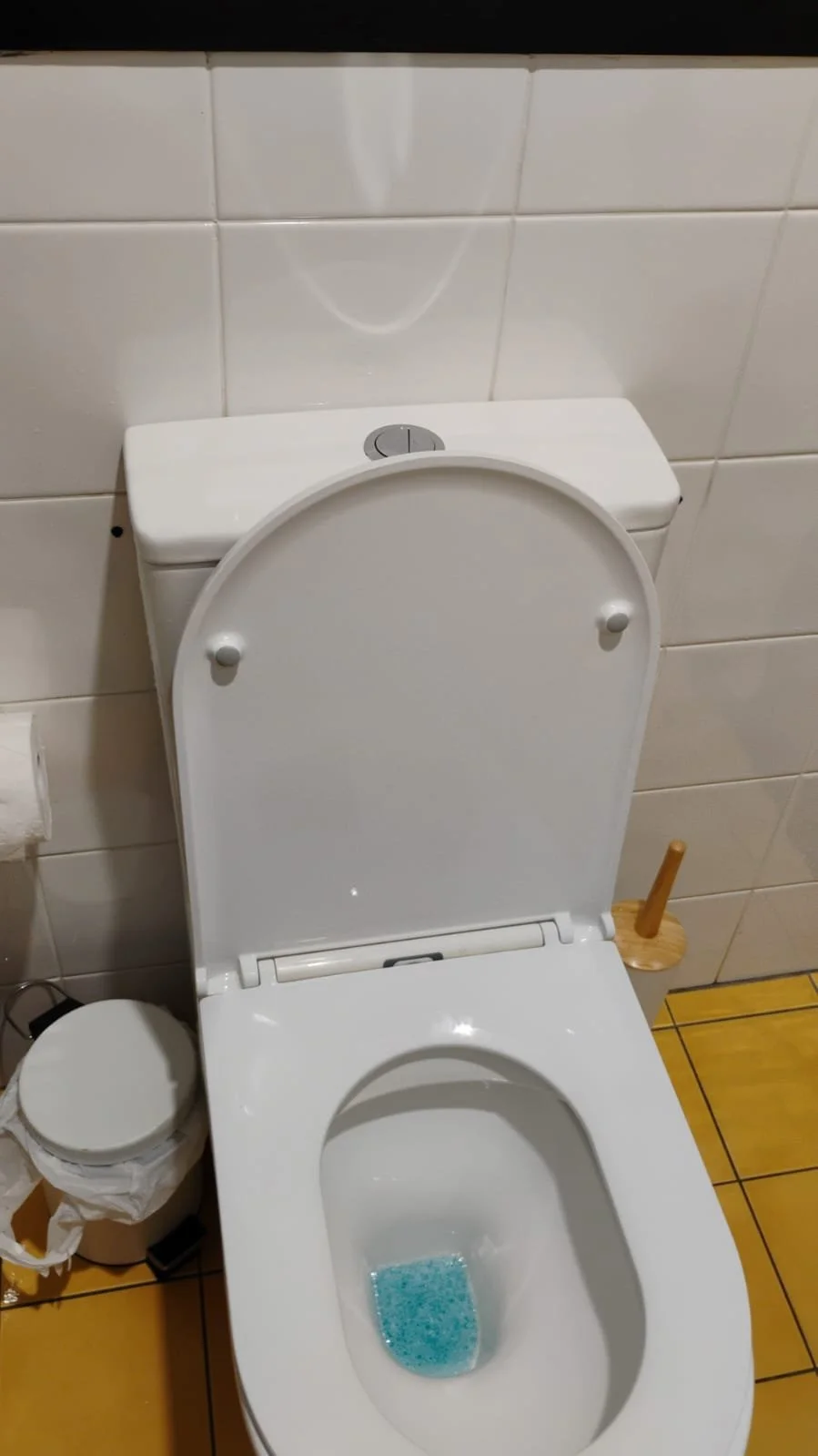 Close-up of a modern white toilet with the lid open, showing blue cleaning tablets inside the bowl. The toilet is against a tiled wall in a bathroom, with a wooden toilet brush and a trash bin nearby.