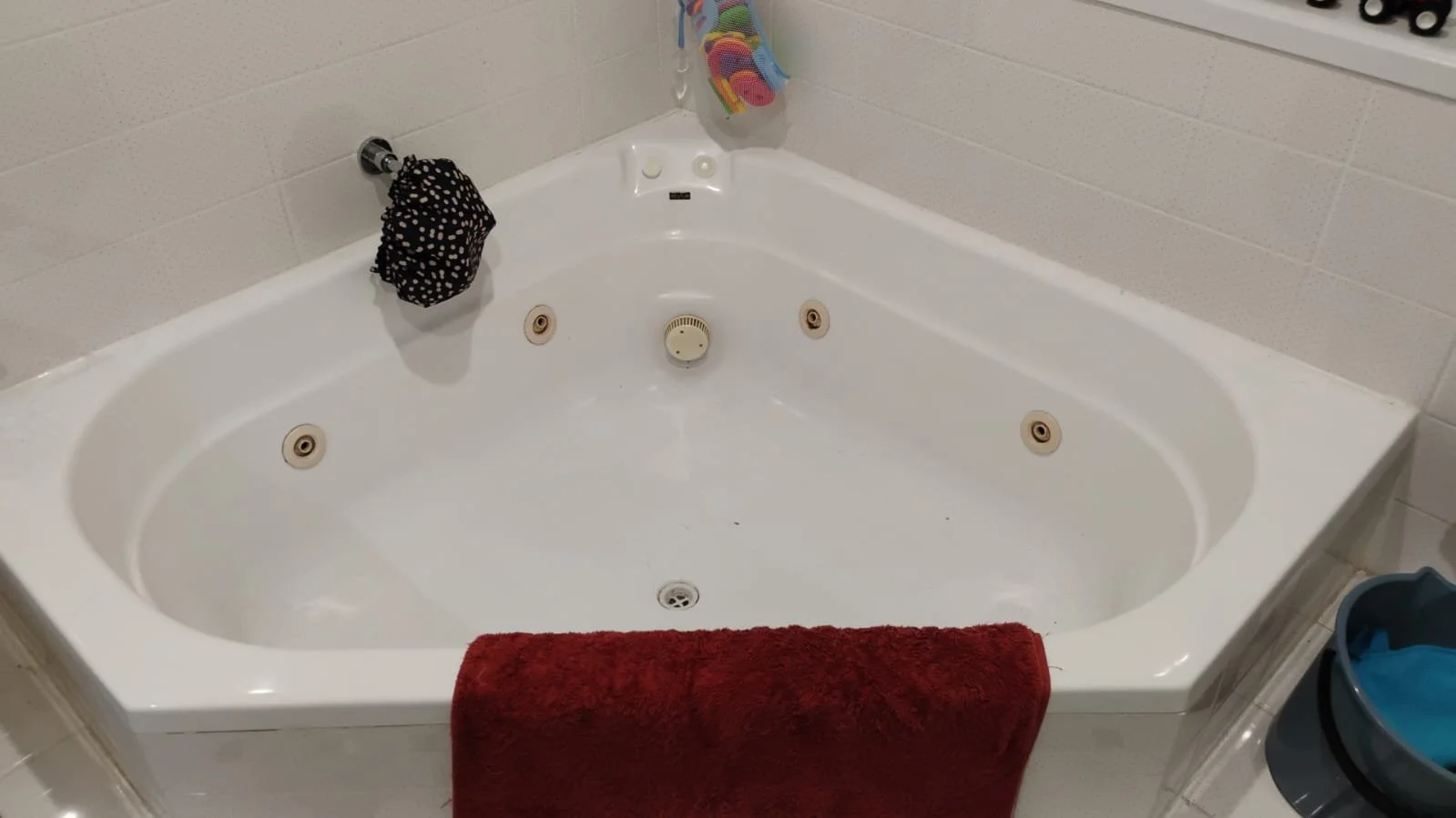 A white corner whirlpool bathtub with six water jets, a red towel draped over the edge, and a black patterned scrubber hanging on a side fixture. The bathtub is in a bathroom with white wall tiles.