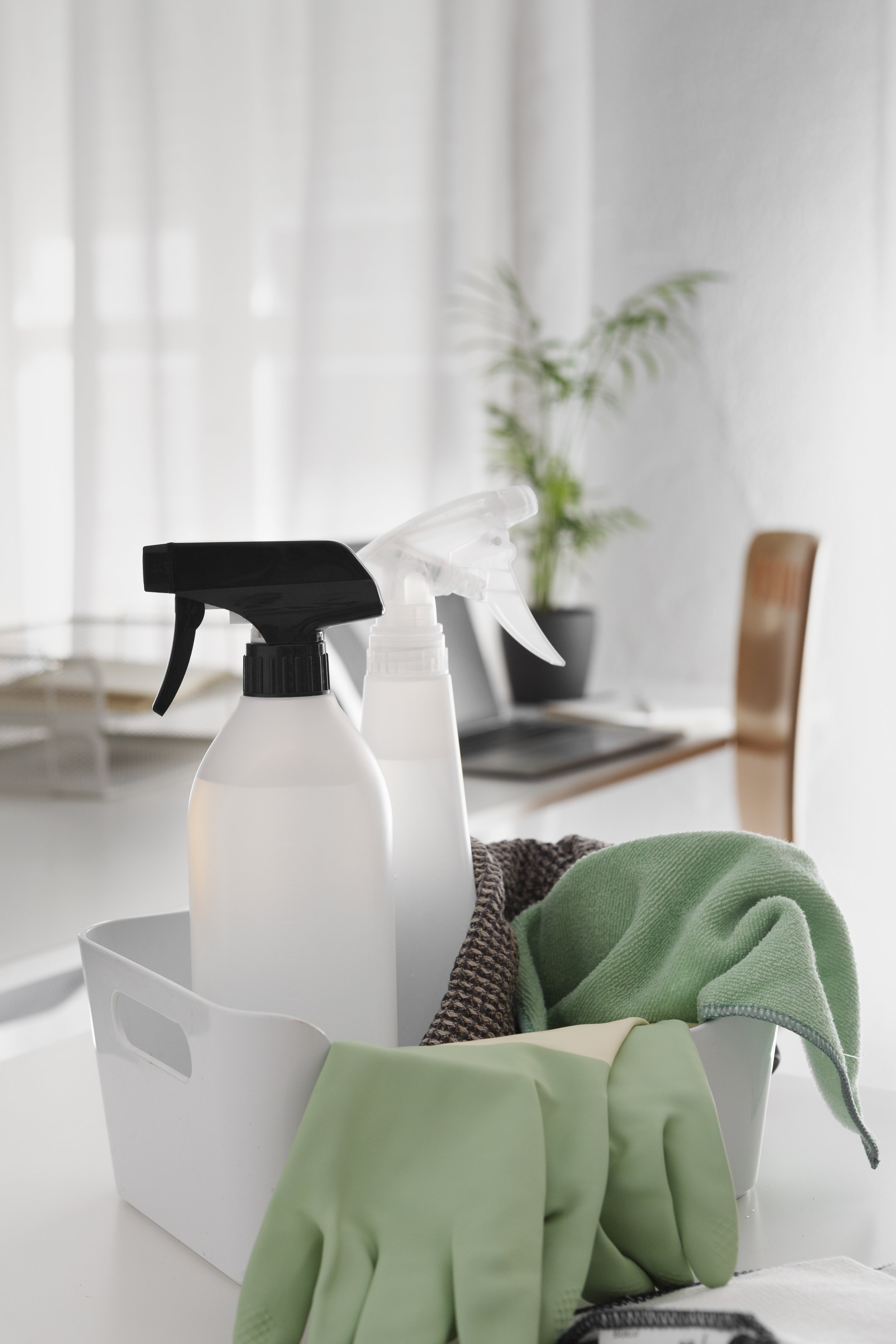 Cleaning supplies on a white container including spray bottles, rubber gloves, and cloths in a sunlit room with a potted plant and a wooden chair in the background.
