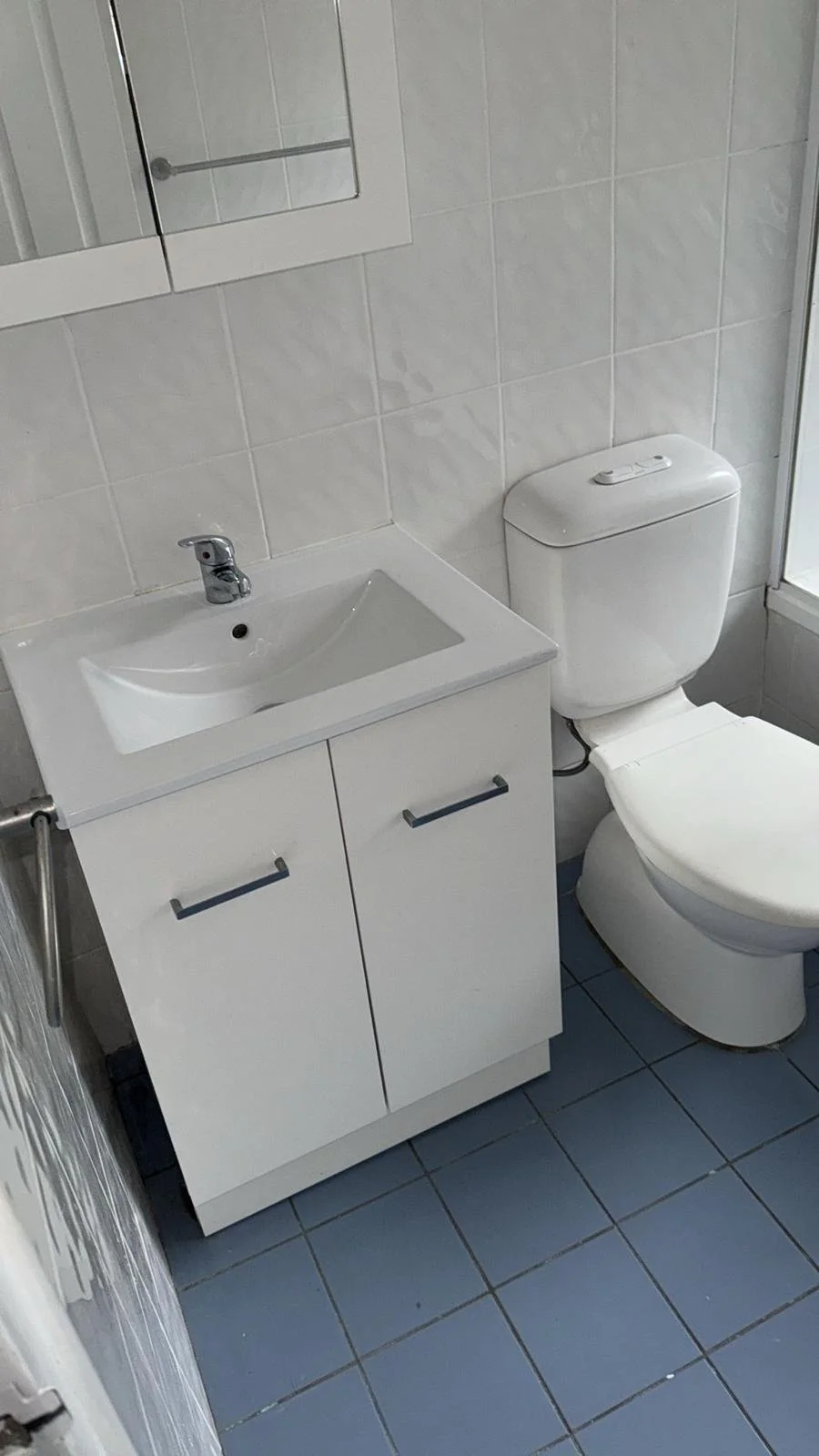 A small bathroom with a white sink and cabinet, a white toilet, gray tiled floor, and tiled walls, with a mirror above the sink.