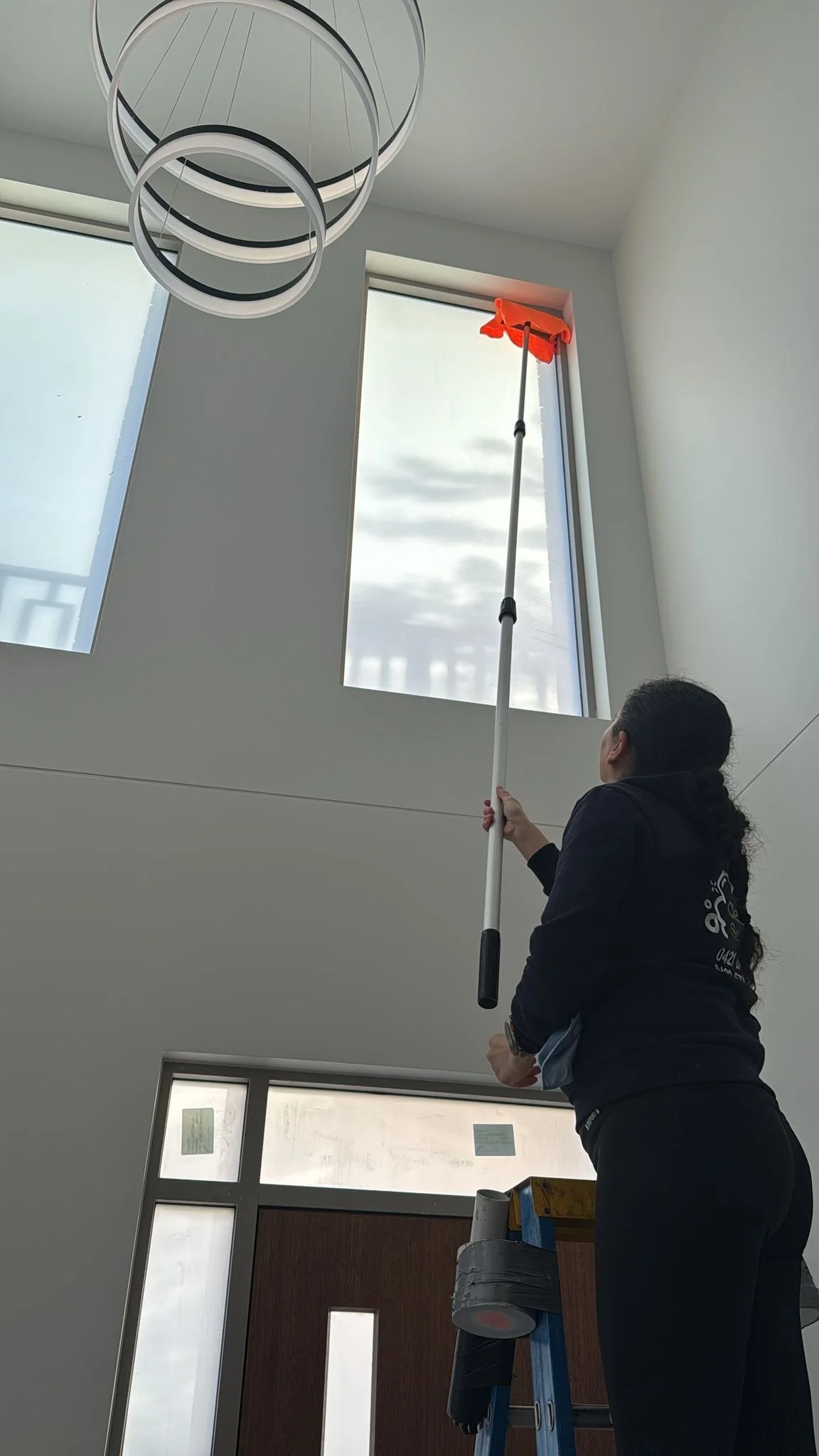 A woman cleaning high windows using an extendable squeegee while standing on a ladder inside a modern building with high ceilings and large windows.