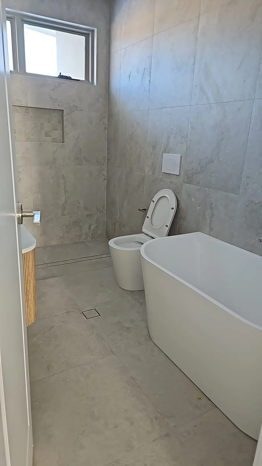 Modern bathroom with a large bathtub, a toilet, a small sink, a small window, and neutral-colored tiles on the walls and floor.