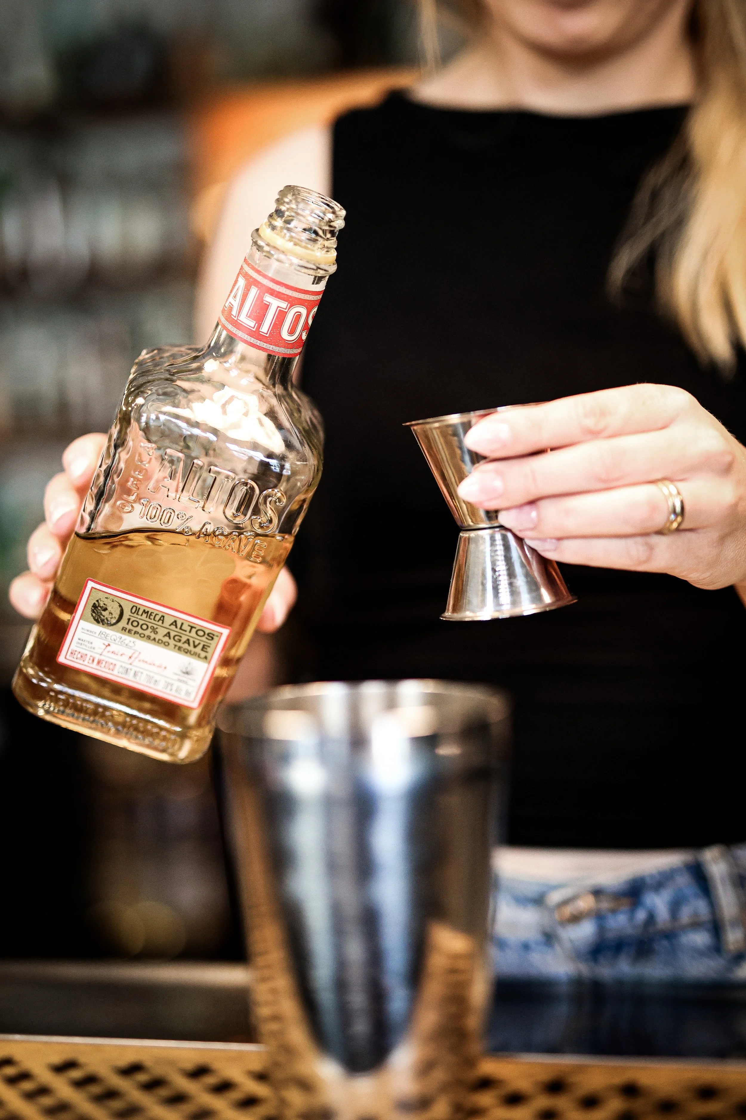 Bartender i en bar, der hælder to forskellige cocktails i glas, med hylder fulde af spirits i baggrunden.