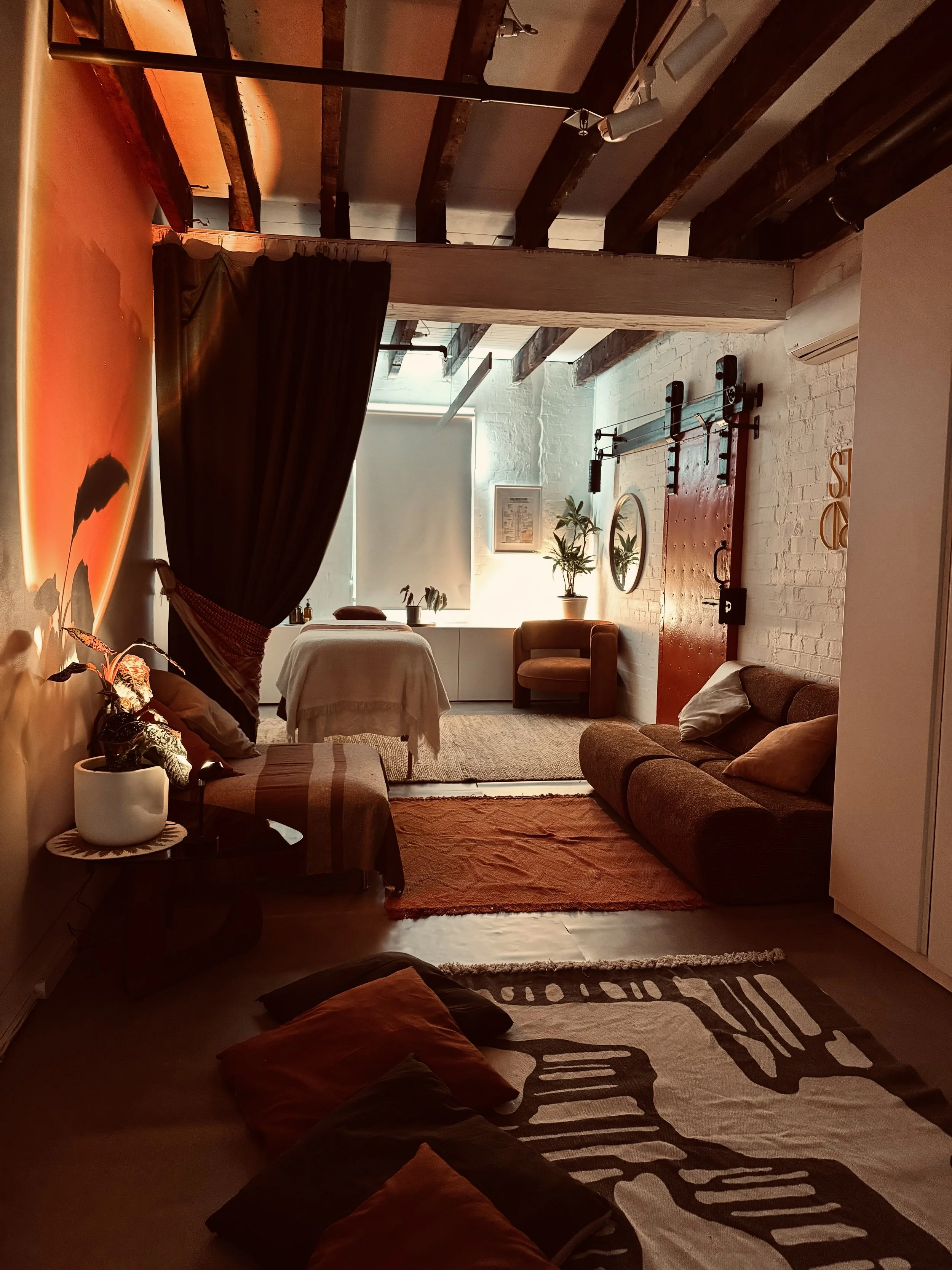 A cosy room with wooden beams on the ceiling, white brick walls, and a mix of furniture including a bed, a brown sofa, a yellow armchair, and various plants. The room is decorated with warm lighting, a rug, and art. A calm space to reconnect.
