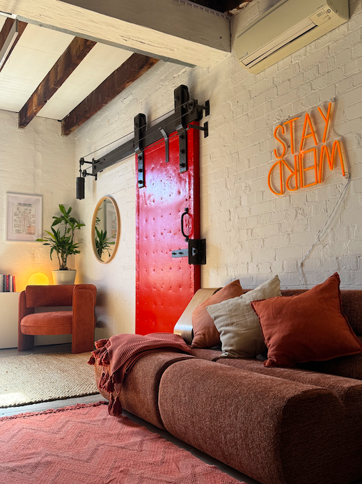 Cosy therapy room featuring a red sliding barn door, a brown couch with throw pillows, a potted plant, a mirror, a neon sign that reads 'STAY WEIRD' and a small lamp on a side table.