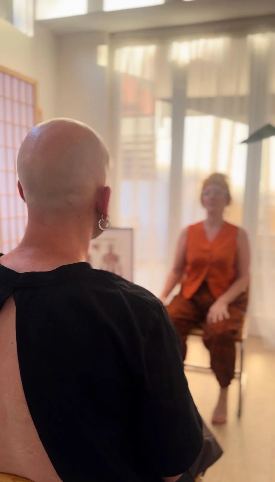 Bald person with earrings sitting and facing a woman in an orange shirt in a bright room with natural light having a somatic therapy session