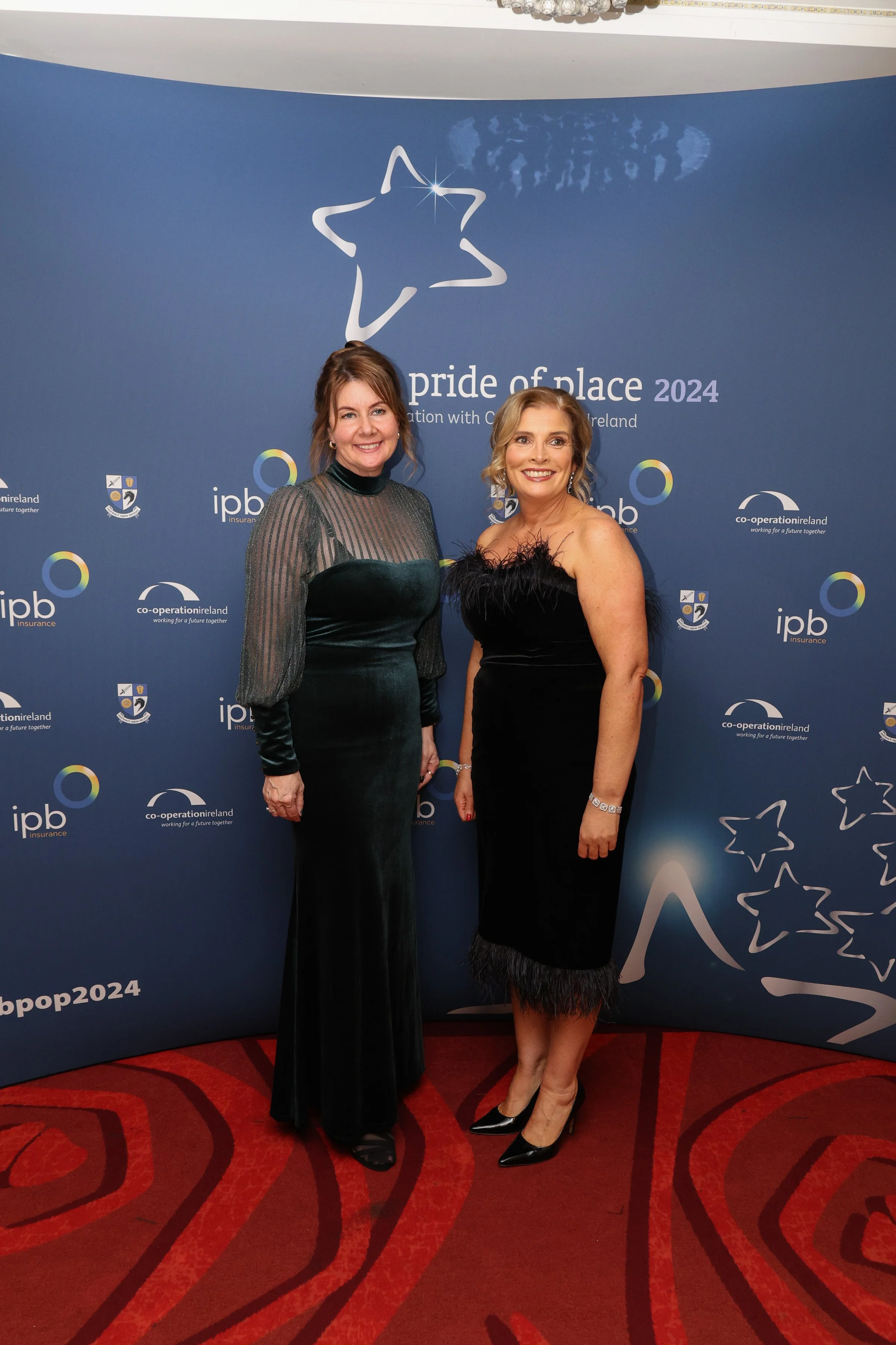 Two women in formal dresses posing in front of a "Pride of Place 2024" event backdrop.