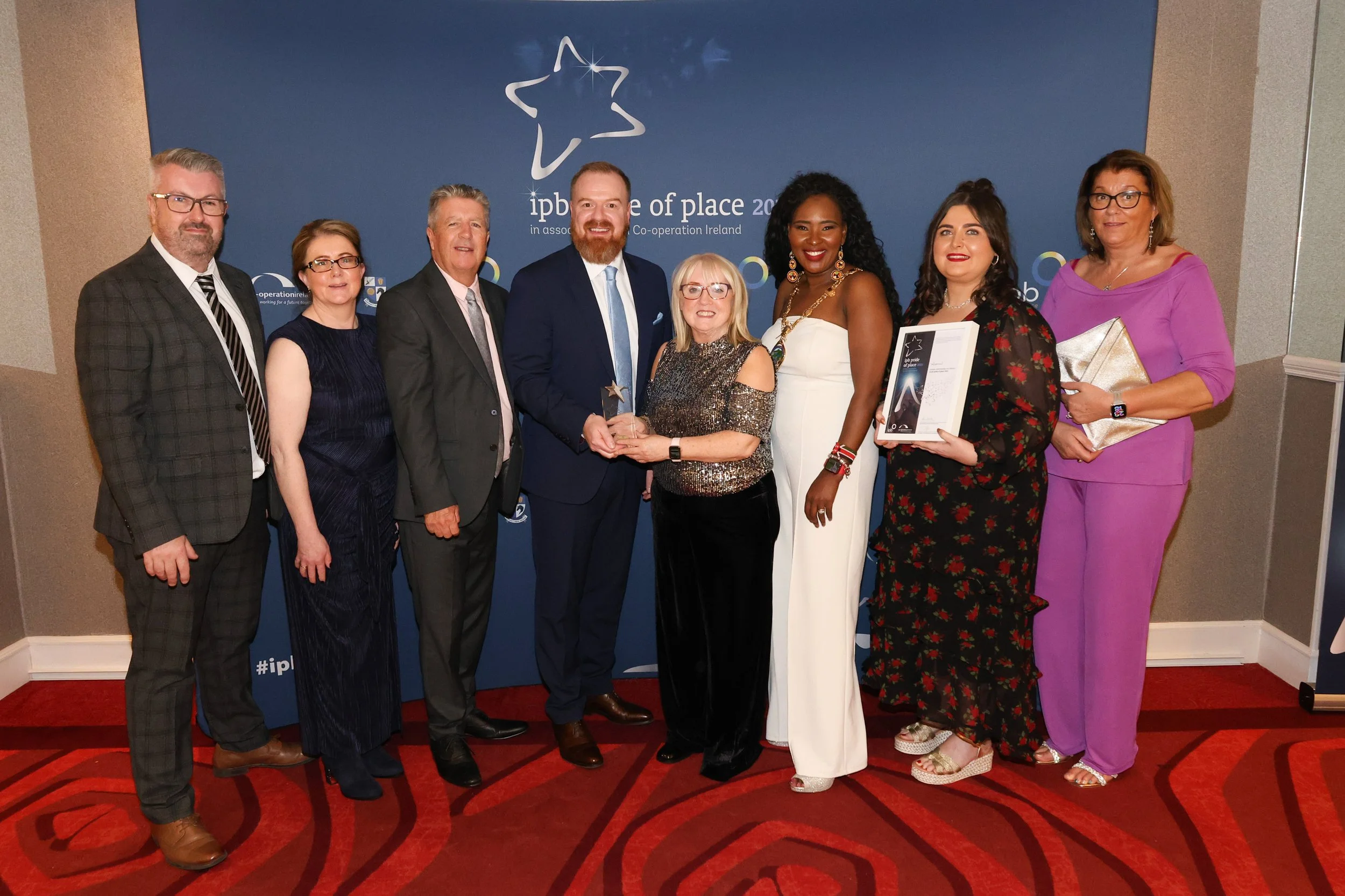 Group of eight people posing for an award ceremony, with a blue backdrop displaying "ipb pride of place 2023."