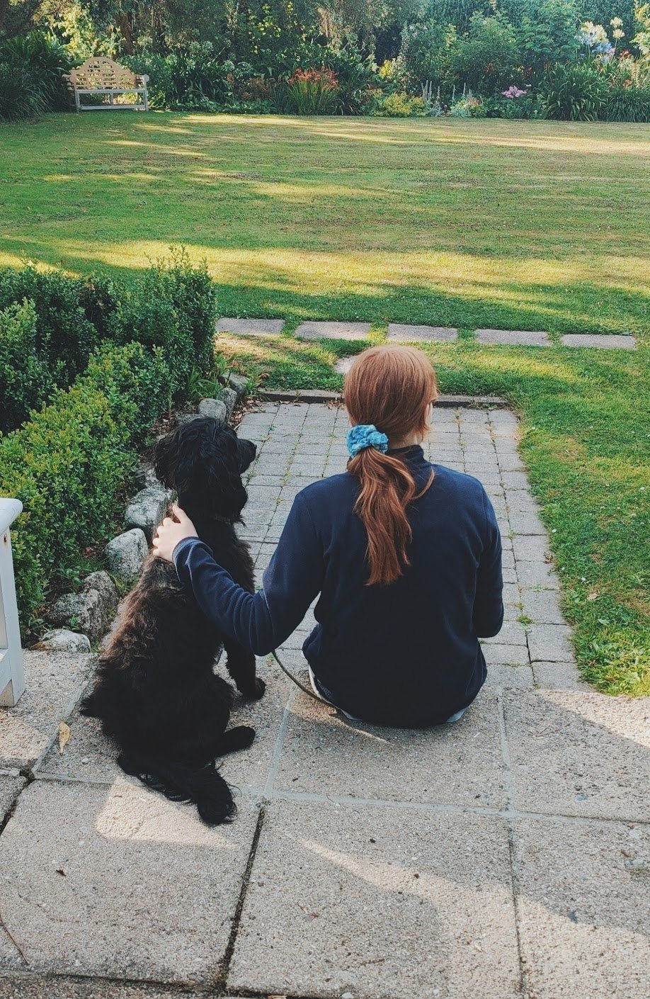 A woman with red hair tied in a ponytail with a blue scrunchie sitting on a stone patio next to a black dog, both looking out at a well-manicured backyard with a lush lawn and garden in the distance.