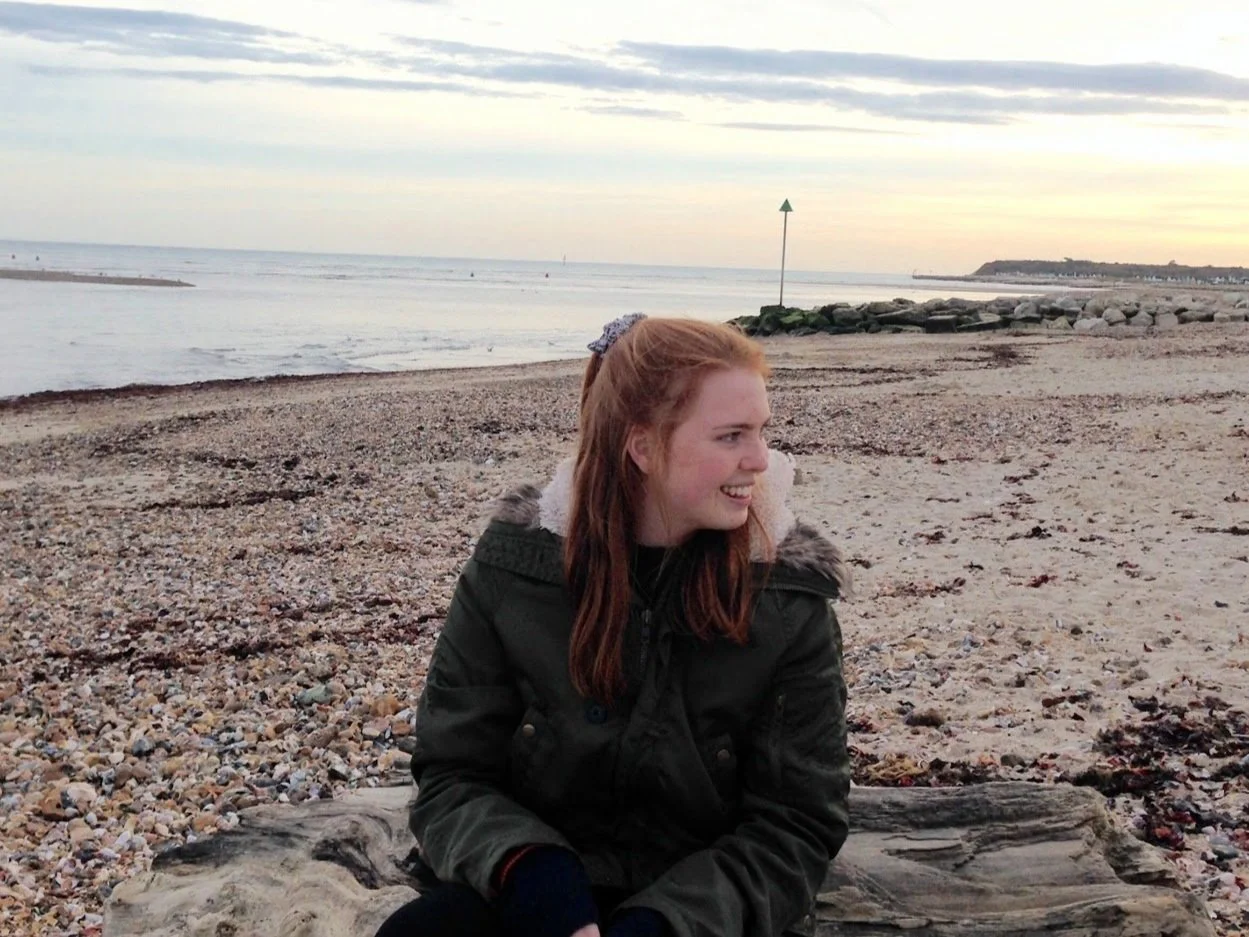 A young woman with red hair sitting on a driftwood log on a pebbled beach, smiling and looking to her right, with the ocean and a cloudy sky in the background.