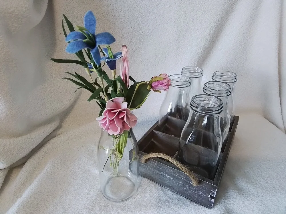 A glass vase with artificial pink and blue flowers next to a wooden tray holding empty glass bottles.