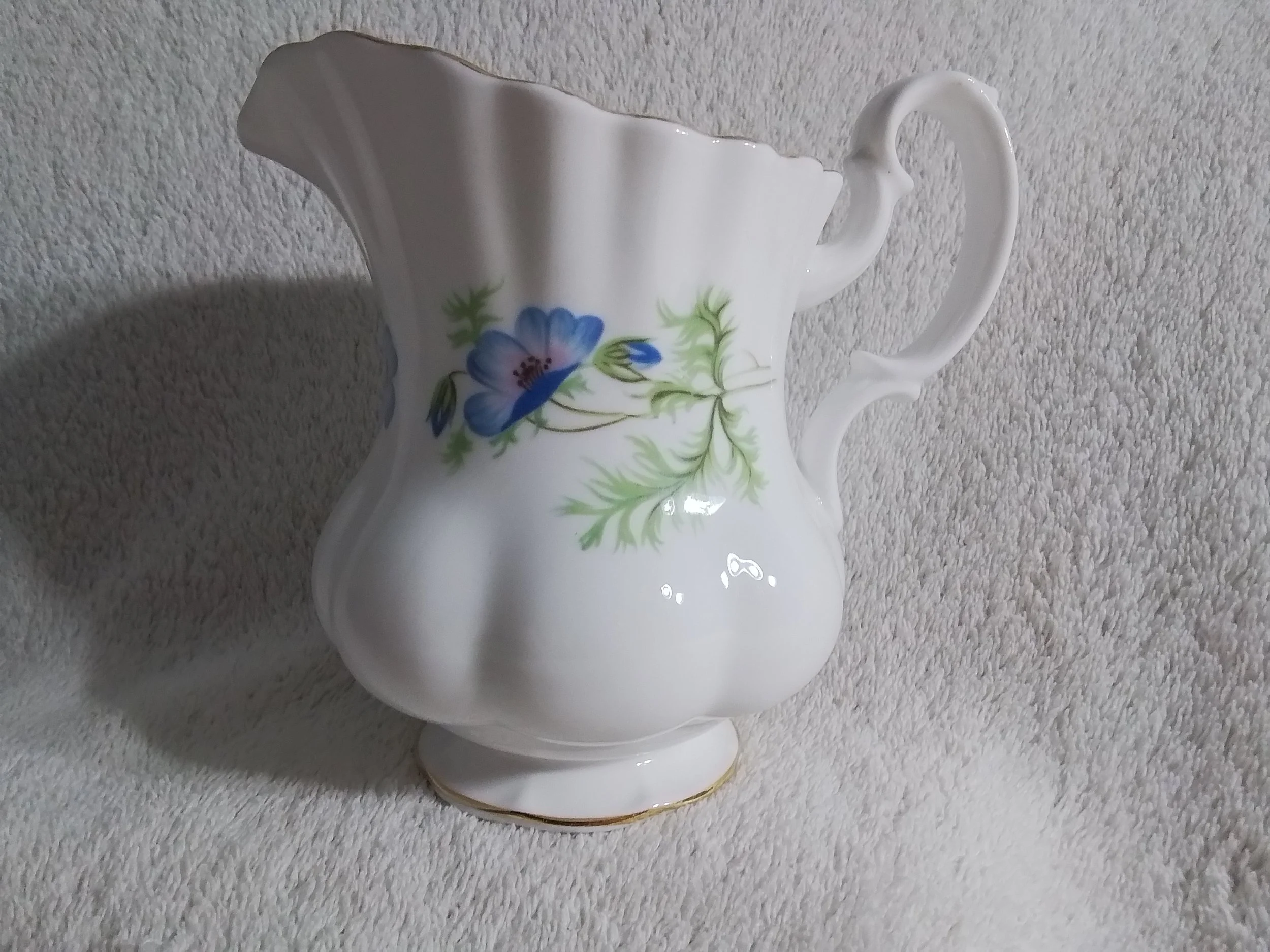 A white ceramic pitcher with hand-painted blue flowers and green leaves, featuring a scalloped rim and a curved handle, placed on a textured off-white surface.