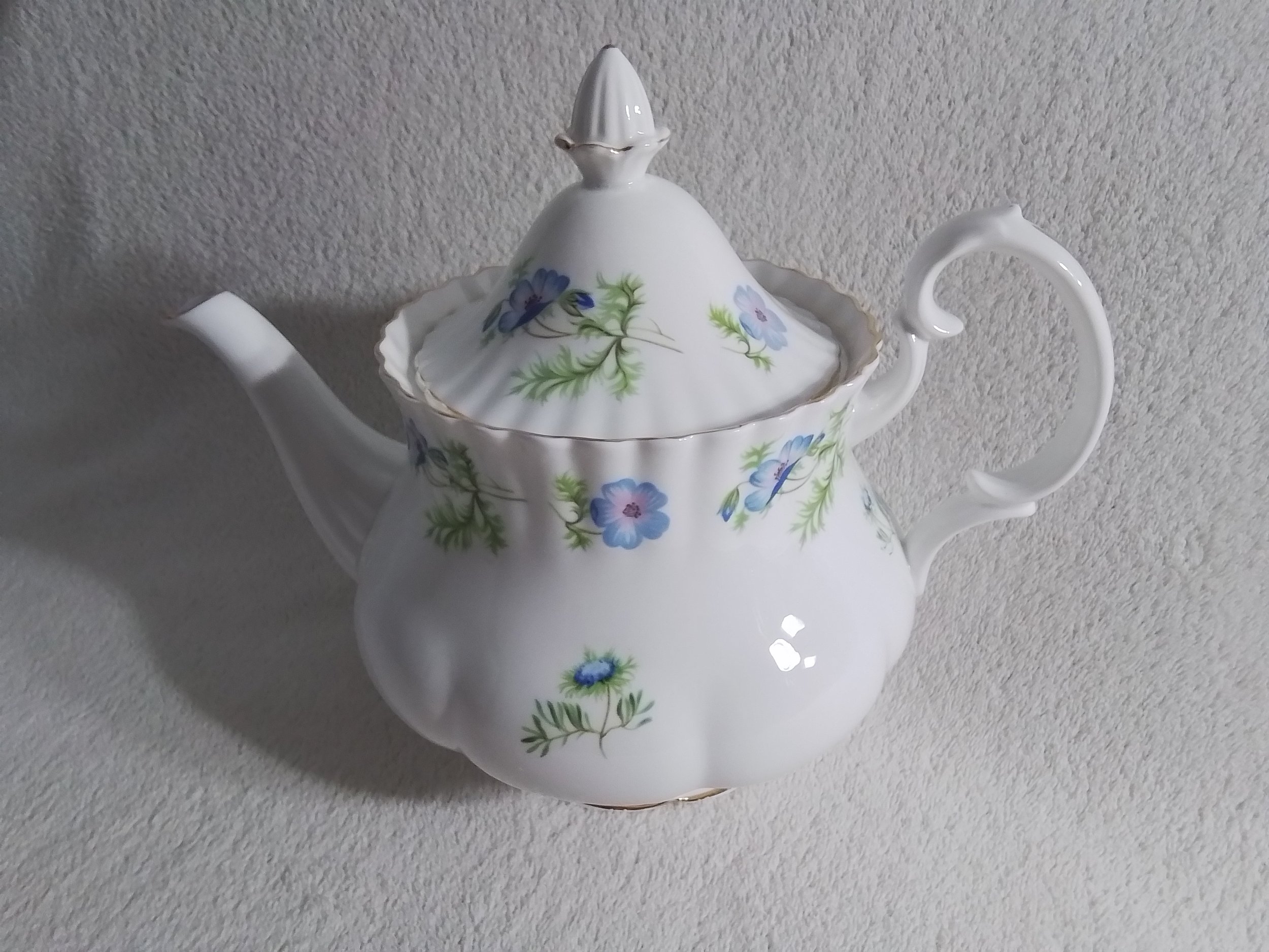 A decorative porcelain teapot with a white floral design, gold accents, and a lid, resting on a light-colored textured surface.