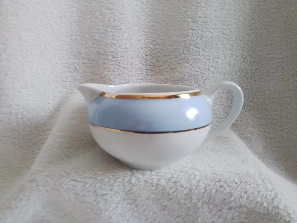 White ceramic creamer with light blue band and gold trim, placed on a soft, textured white surface with a matching textured background.