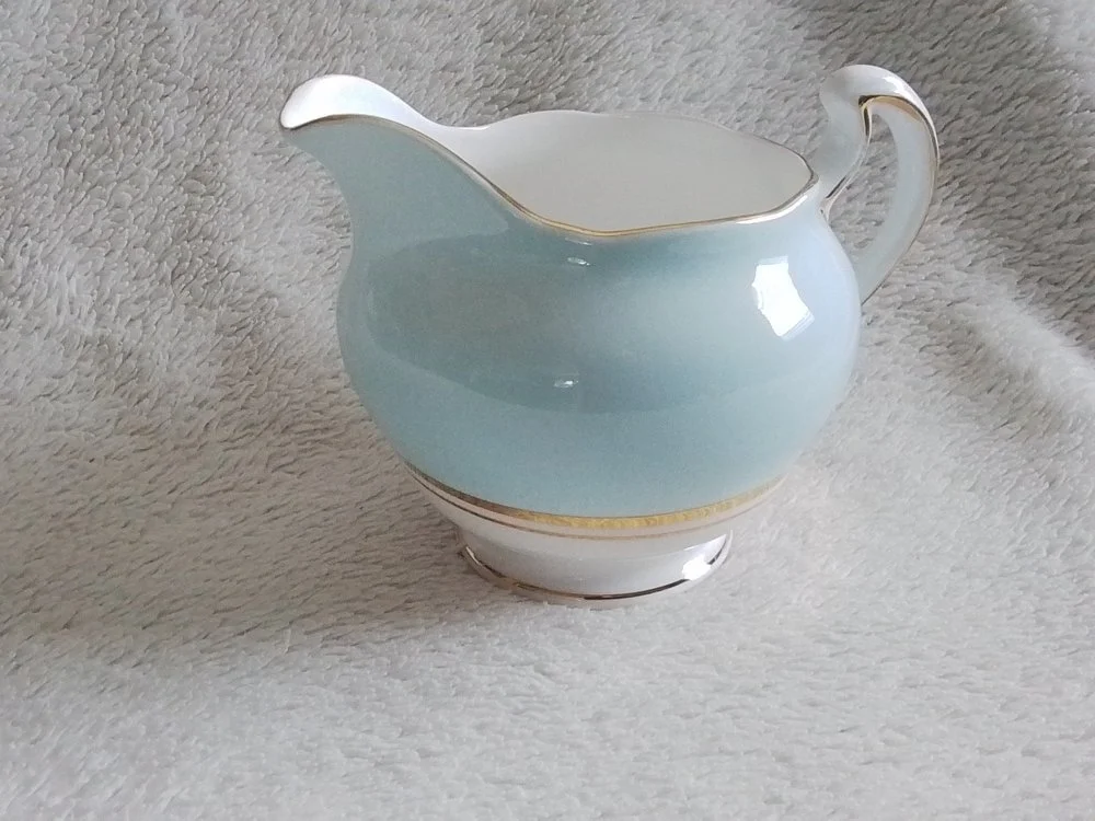 A light blue and white porcelain creamer with gold trim, placed on a soft, light-colored textured fabric.