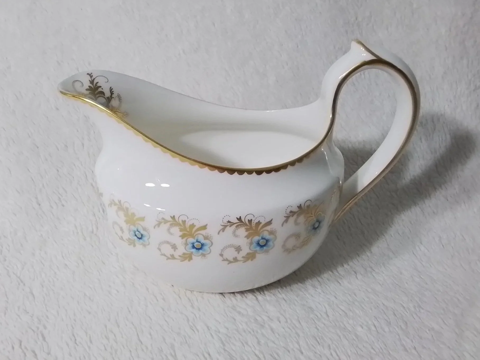 A white porcelain gravy boat with gold trim and a floral pattern of blue flowers and gold leaves.
