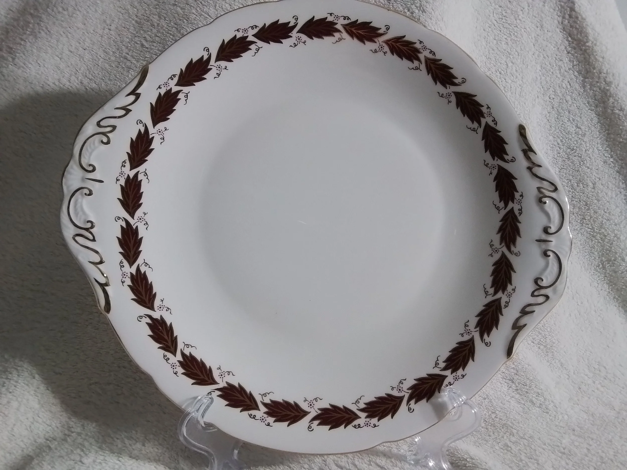 Decorative ceramic plate with a white background and a brown leaf pattern along the rim, displayed on a clear stand on a white textured surface.