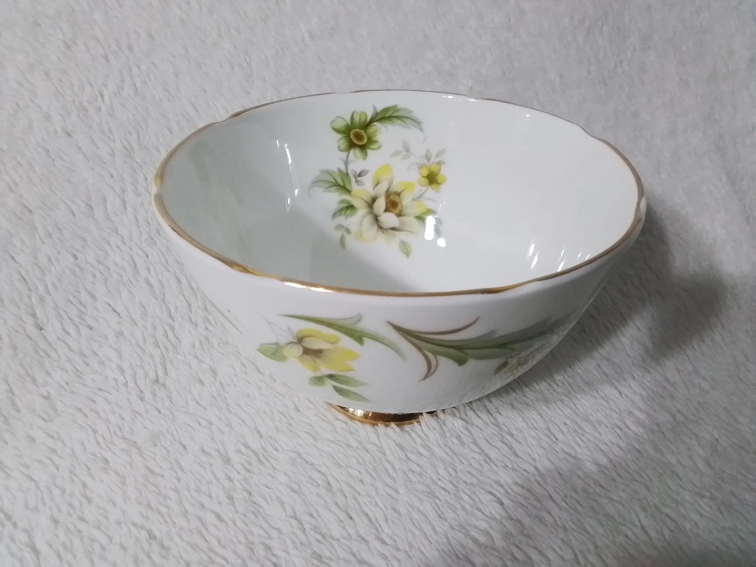 A white porcelain bowl with a floral pattern of yellow and white flowers and green leaves, accented with gold trim. The bowl is placed on a light textured surface.