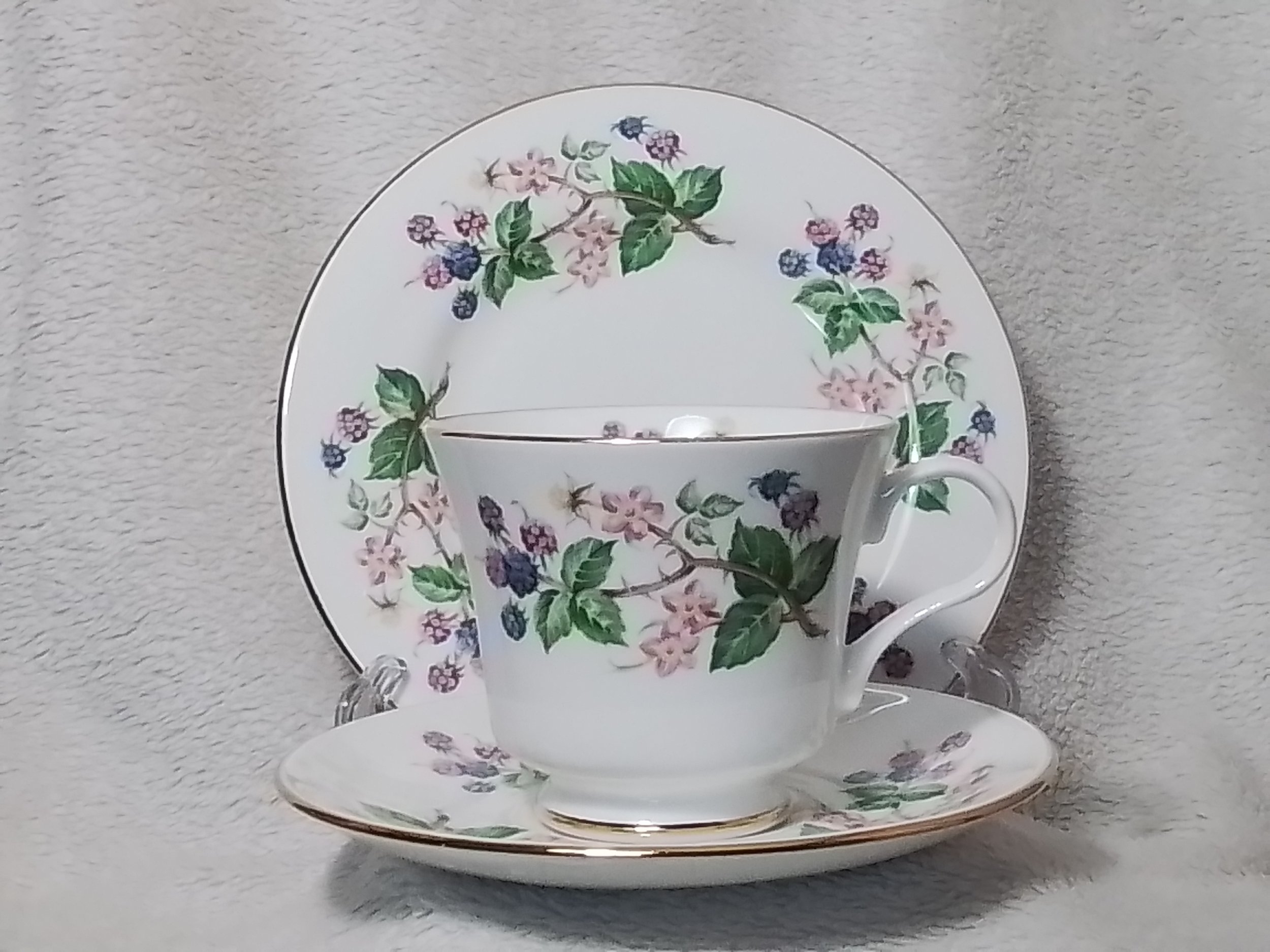 A porcelain tea cup and saucer set with a floral pattern of pink, purple, and blue flowers with green leaves, placed in front of a matching floral plate against a gray fabric background.