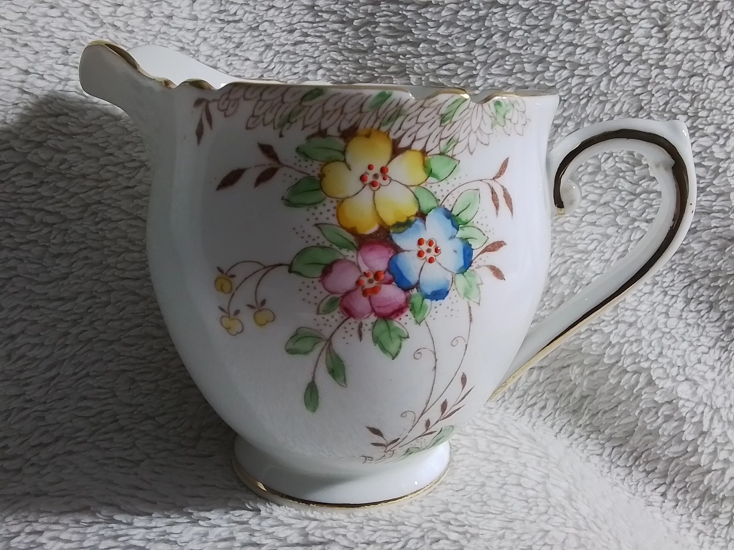 A white porcelain teapot with floral decoration featuring yellow, pink, and blue flowers with green leaves, gold accents, and a handle with gold trim, placed on a textured fabric surface.