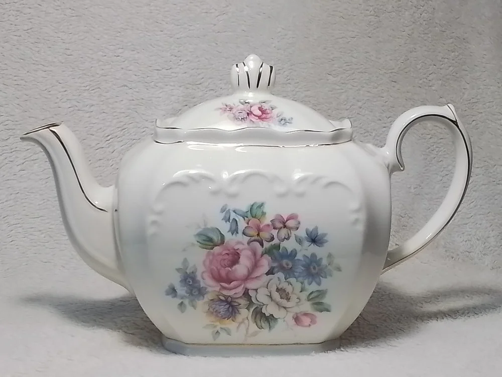 White porcelain teapot with floral pattern and gold accents on the lid, spout, and handle, set against a neutral background.