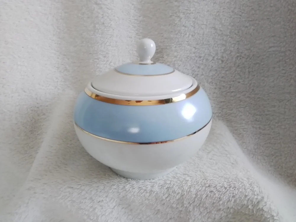 A decorative porcelain jar with a lid, light blue and white color, accented with gold trim, on a soft white surface with a textured background.