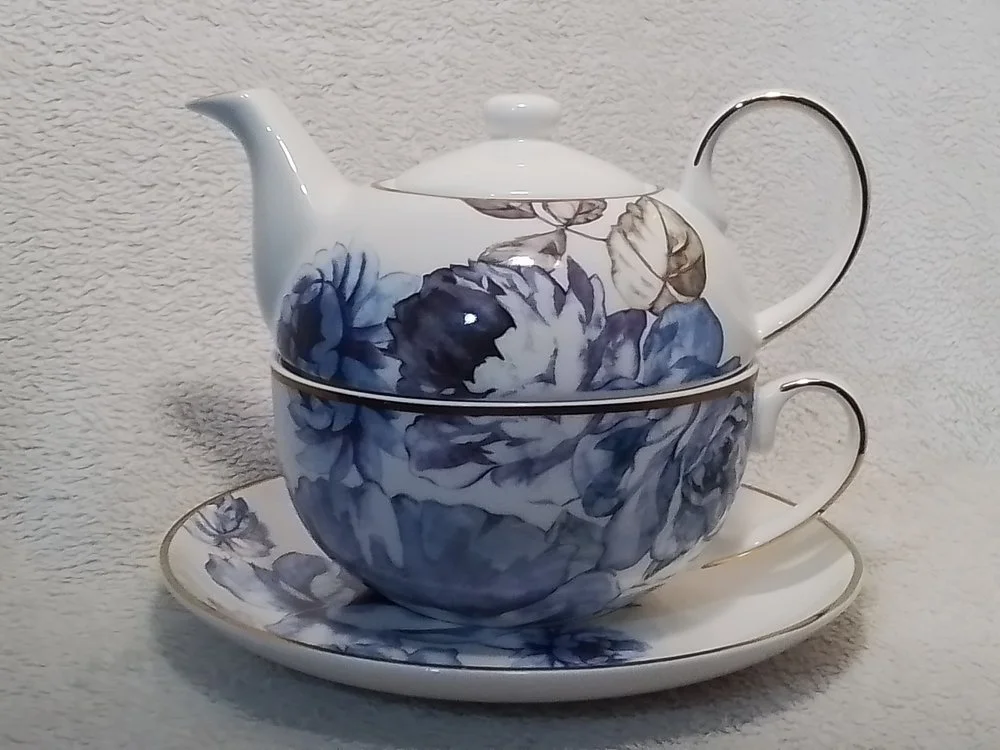 A porcelain tea set with a teapot, a cup, and a saucer, decorated with blue and gray floral patterns, displayed against a textured beige background.