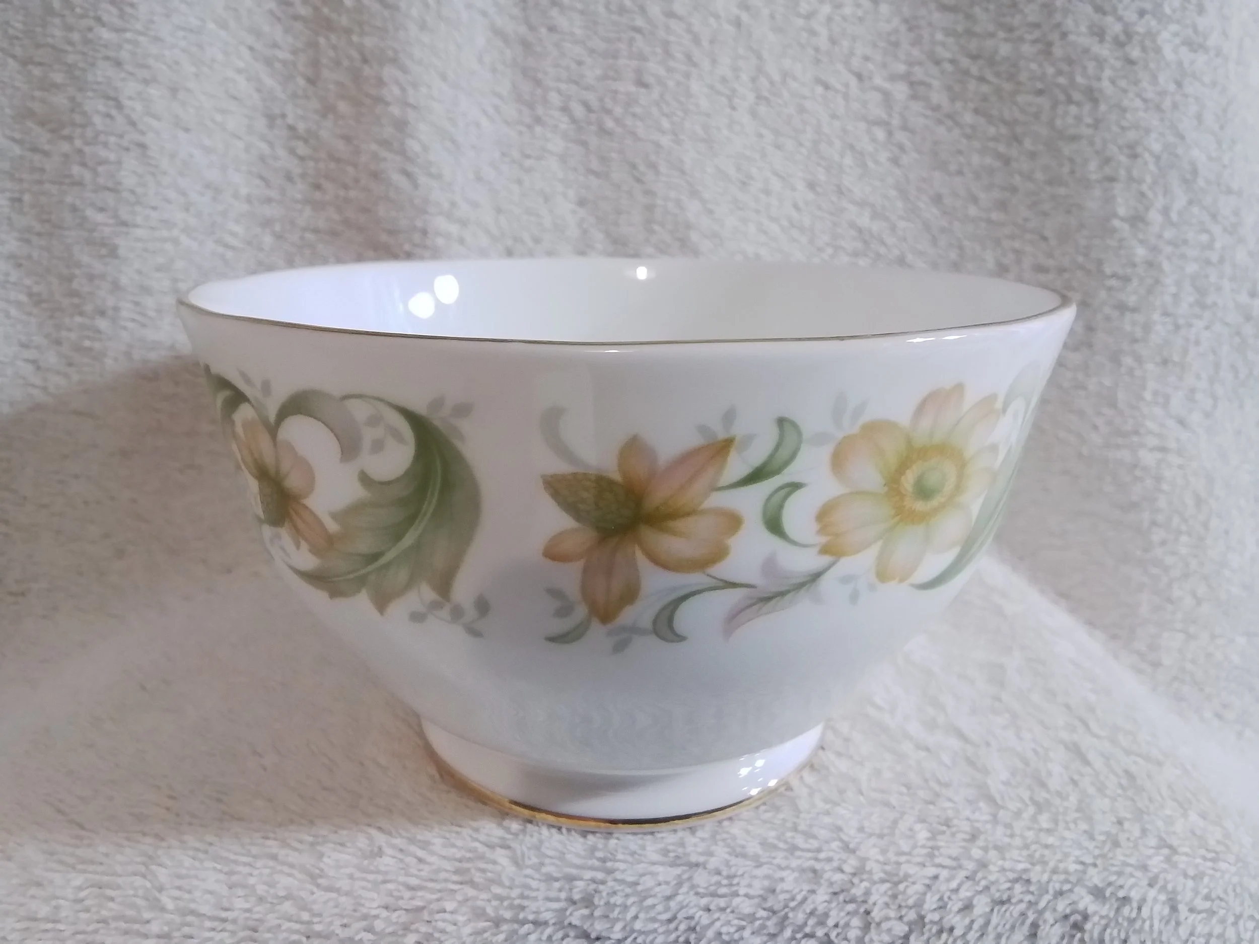 A white ceramic bowl with a gold rim and floral pattern of yellow and beige flowers with green leaves, resting on a white textured surface.