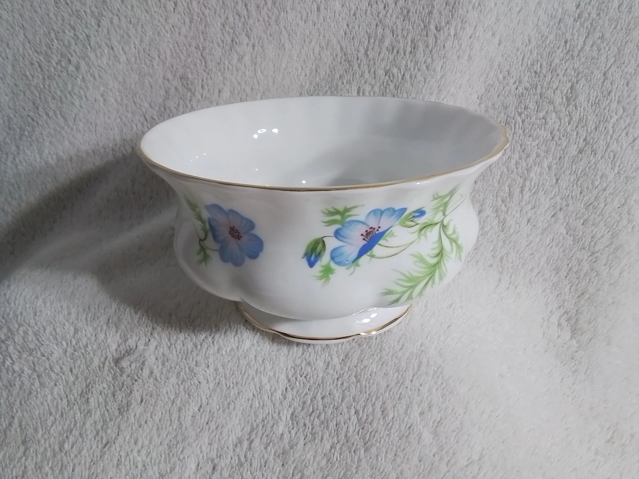 A white porcelain bowl with a floral pattern of blue flowers and green leaves, trimmed with a gold rim, placed on a soft, textured white fabric surface.
