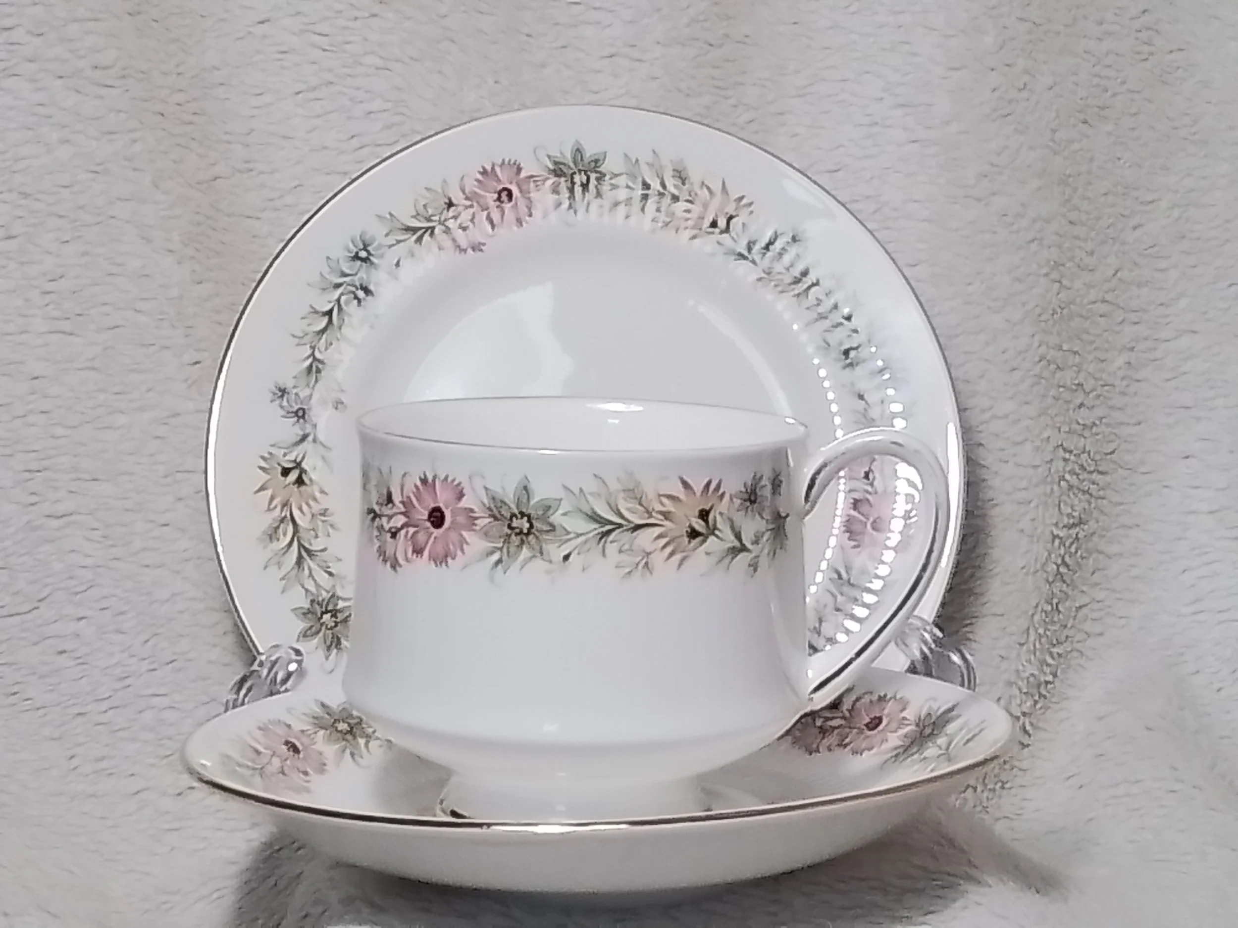 A floral patterned teacup, saucer, and dessert plate with pink, green, and beige flowers on a white background arranged on a textured gray surface.