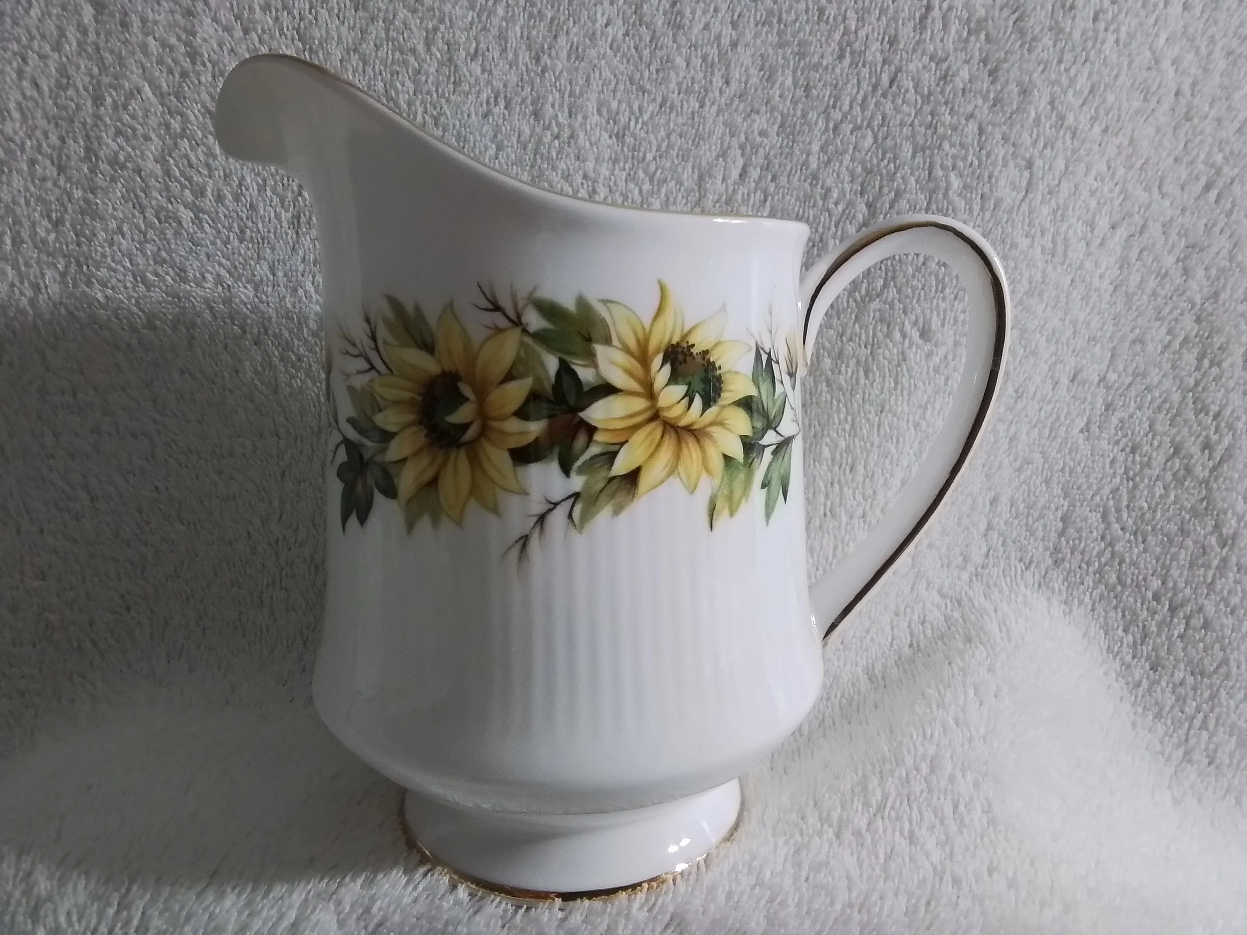 White porcelain pitcher with a floral pattern of yellow sunflowers and green leaves, gold accents on the rim and handle, placed on a textured light gray surface.