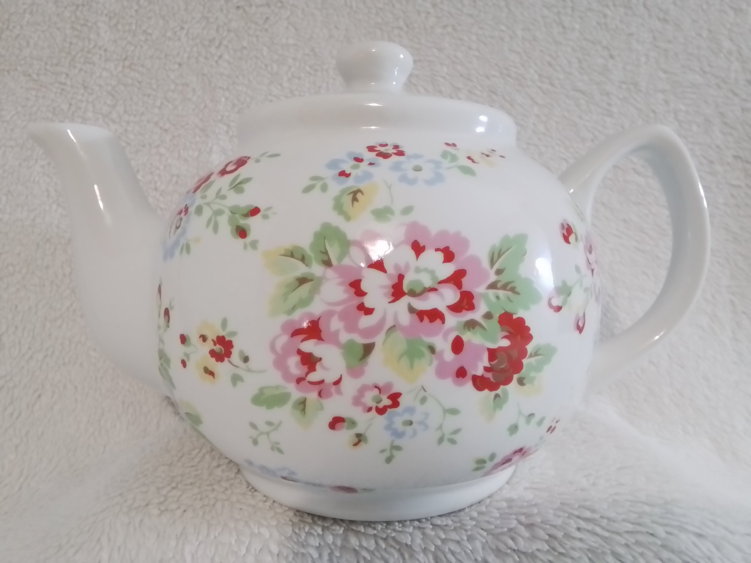 White ceramic teapot with floral pattern of pink, red, blue, and green flowers and leaves, set against a white textured background.