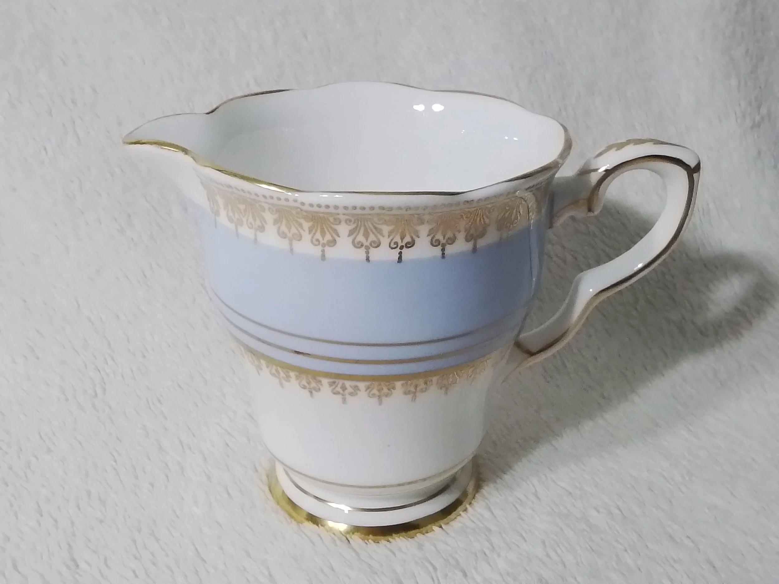 Ceramic creamer with white and gold decorative pattern, on a white textured surface.