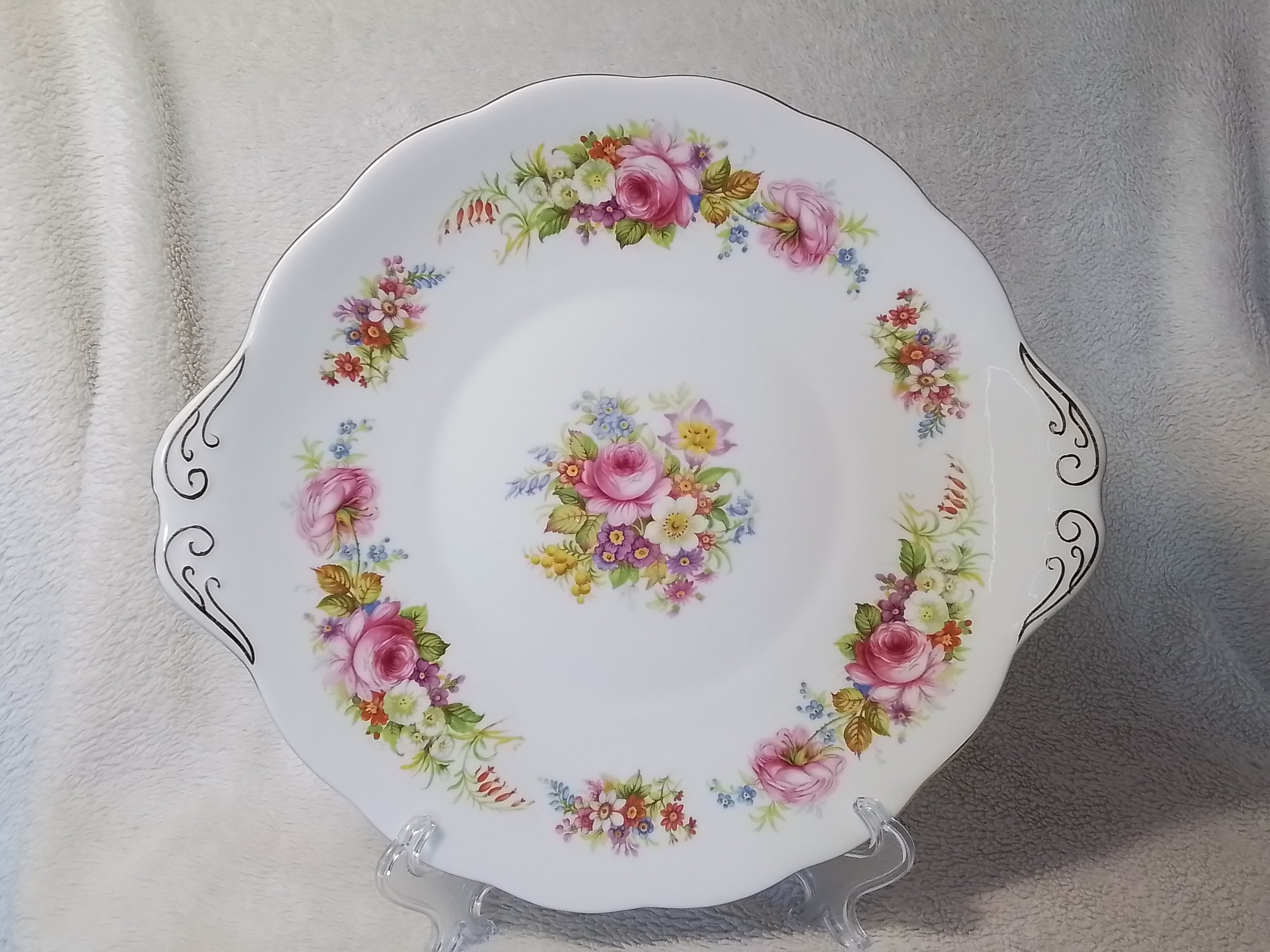 A decorative porcelain plate with a floral pattern, featuring pink roses and other colorful flowers, displayed on a clear stand against a soft fabric background.