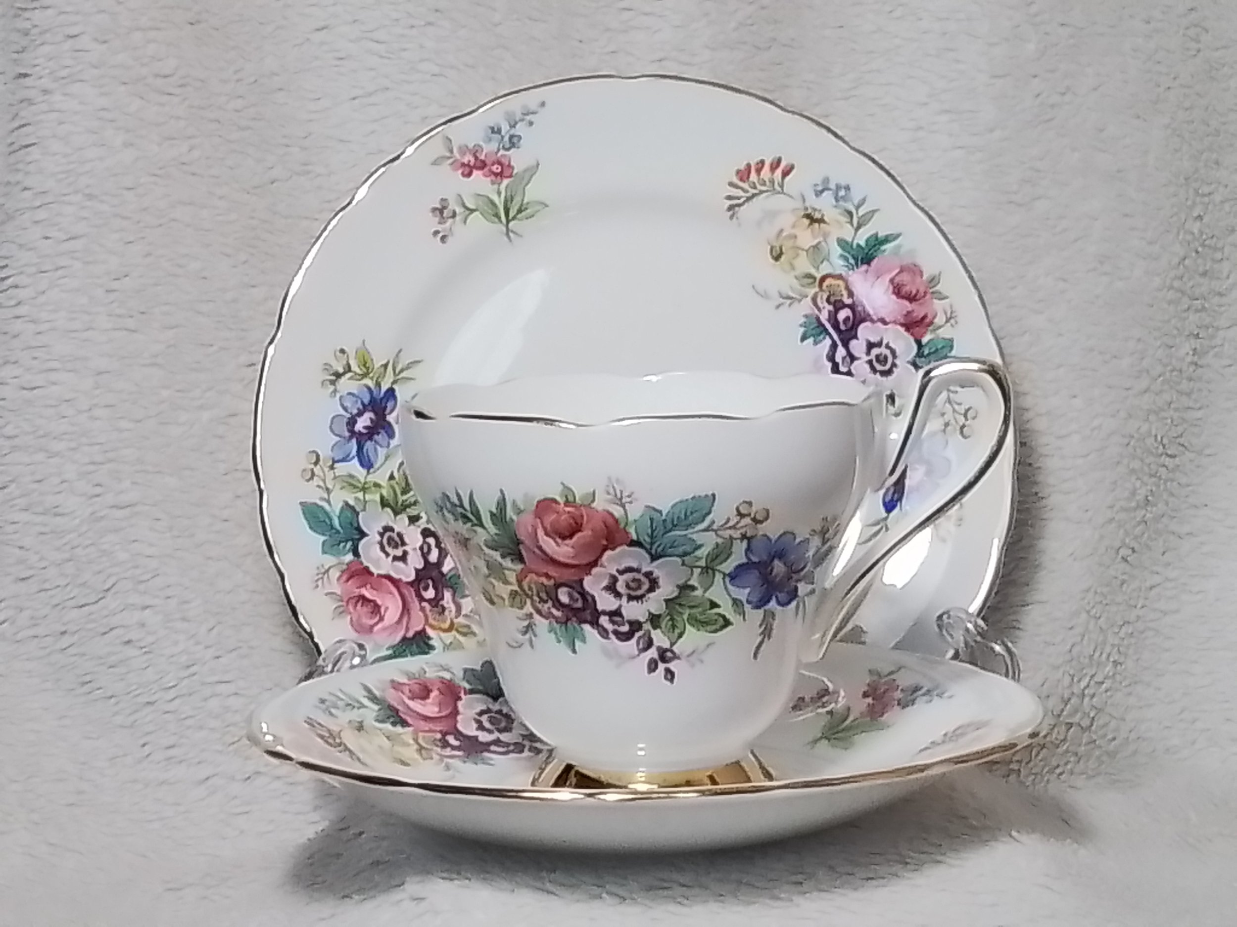 A floral patterned teacup with matching saucer and dessert plate, all with gold trim, placed on a textured grey surface.