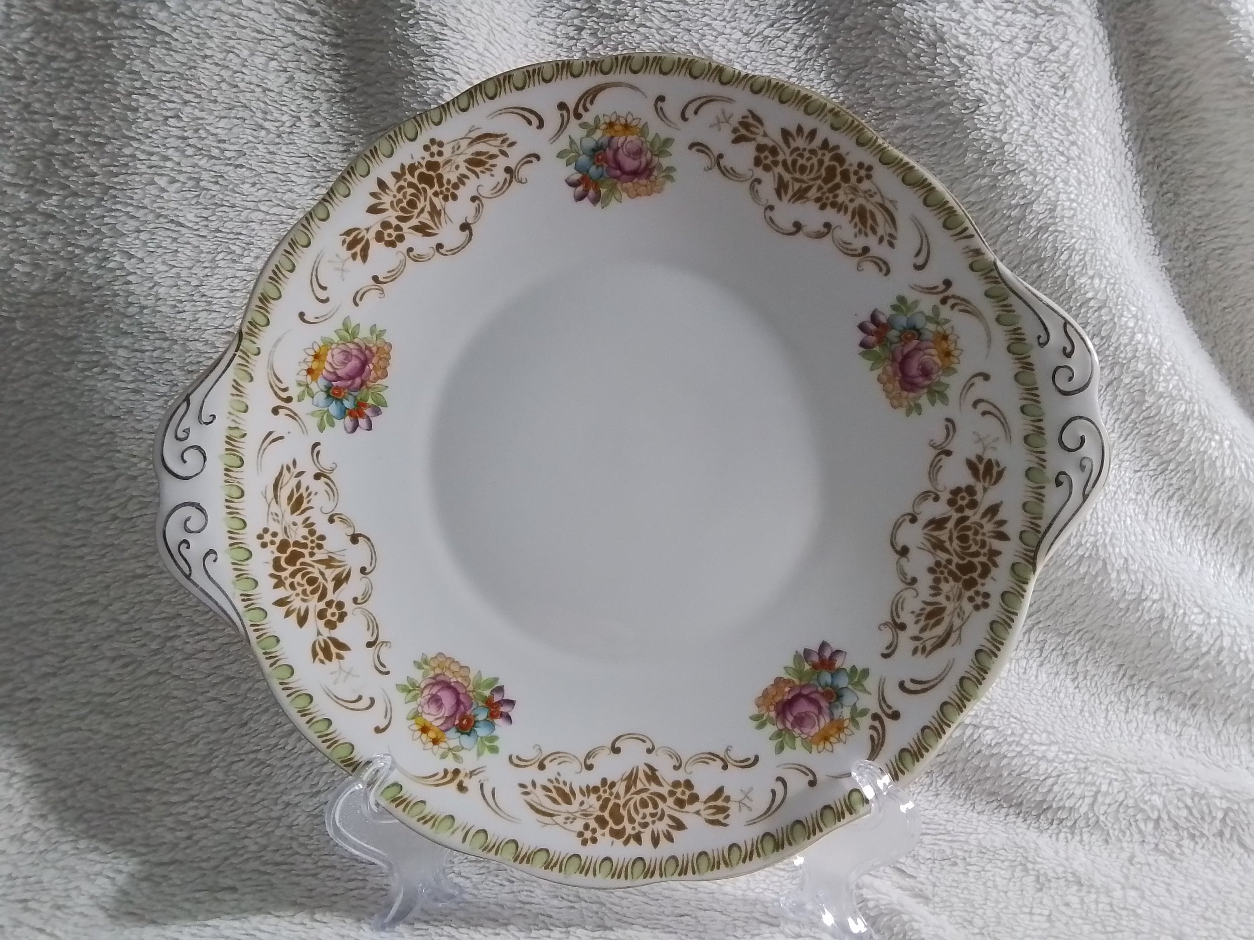 A decorative porcelain plate with floral and gold patterns, displayed on a clear stand on a soft, textured surface.