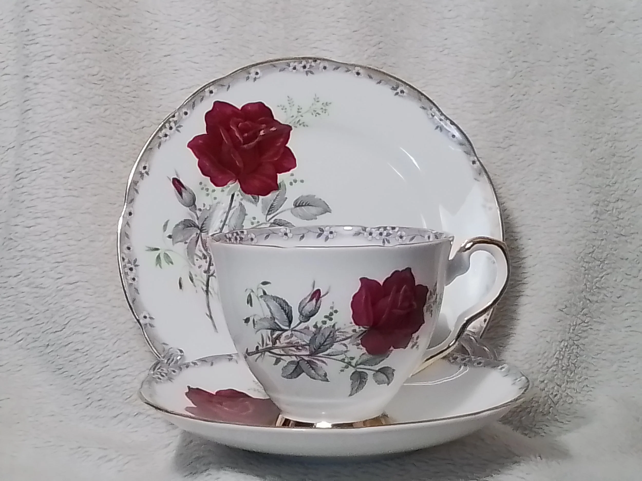 A porcelain teacup and saucer with red roses and green leaves pattern, placed in front of a matching plate with the same floral design, set against a textured light background.