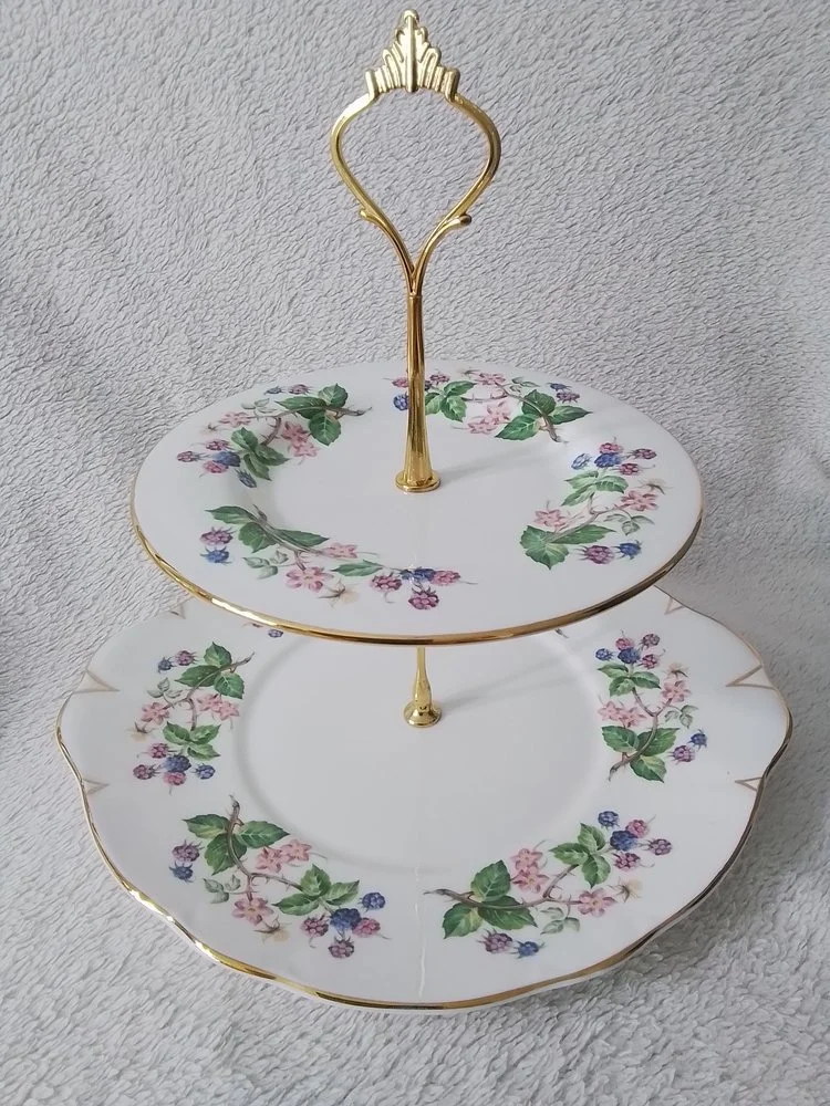 Two-tier floral porcelain serving stand with gold accents against a textured light gray background.