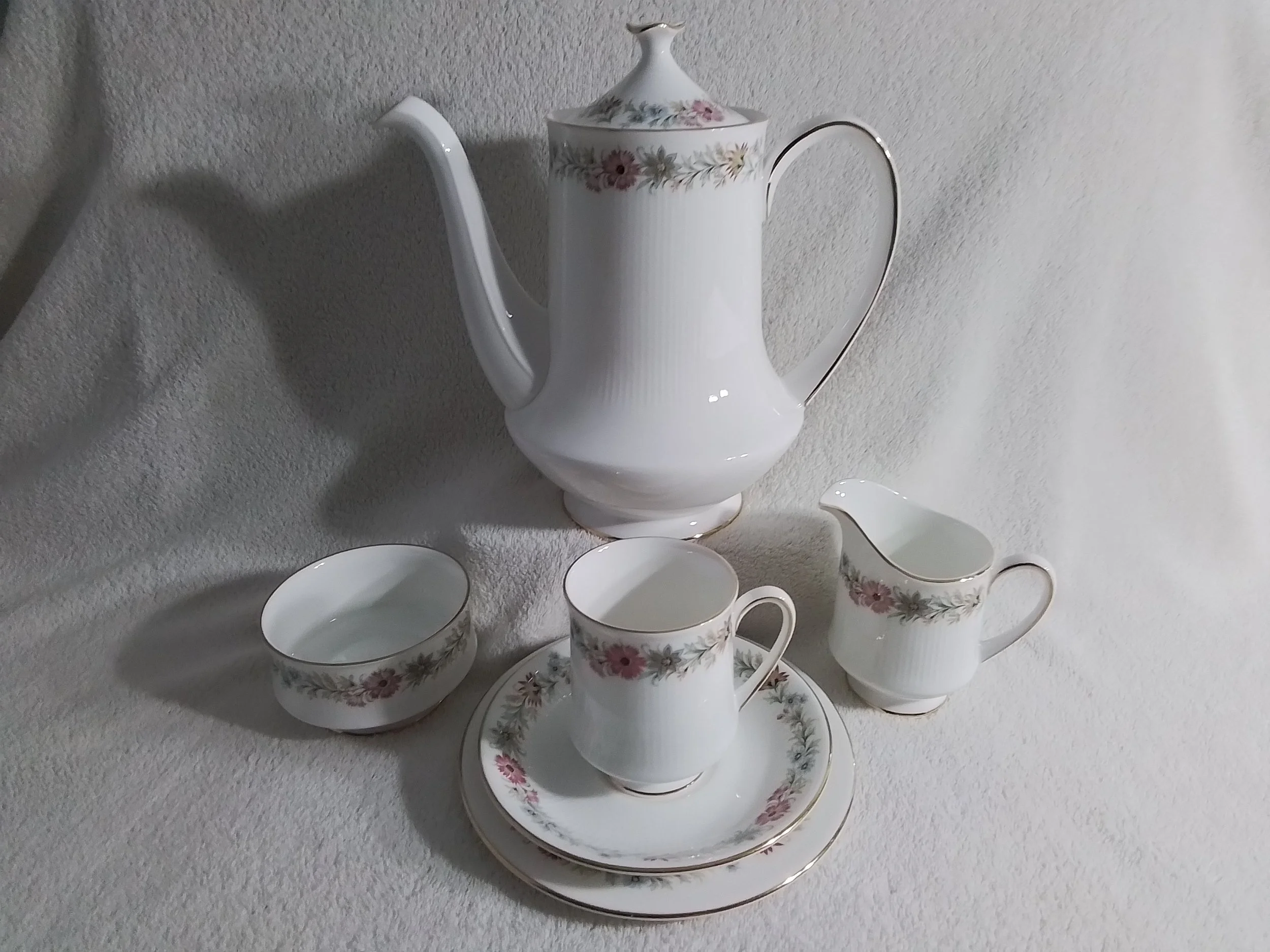 A white porcelain tea set with floral pattern includes a tall teapot, a creamer, two cups on saucers, and a small bowl, all arranged on a light-colored fabric background.